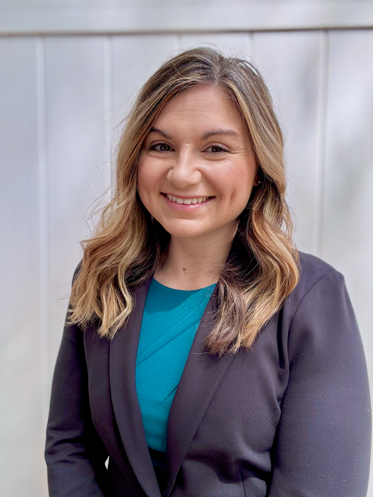 A woman with shoulder-length wavy blonde hair smiling, wearing a teal blouse and a black blazer, standing against a light gray background.