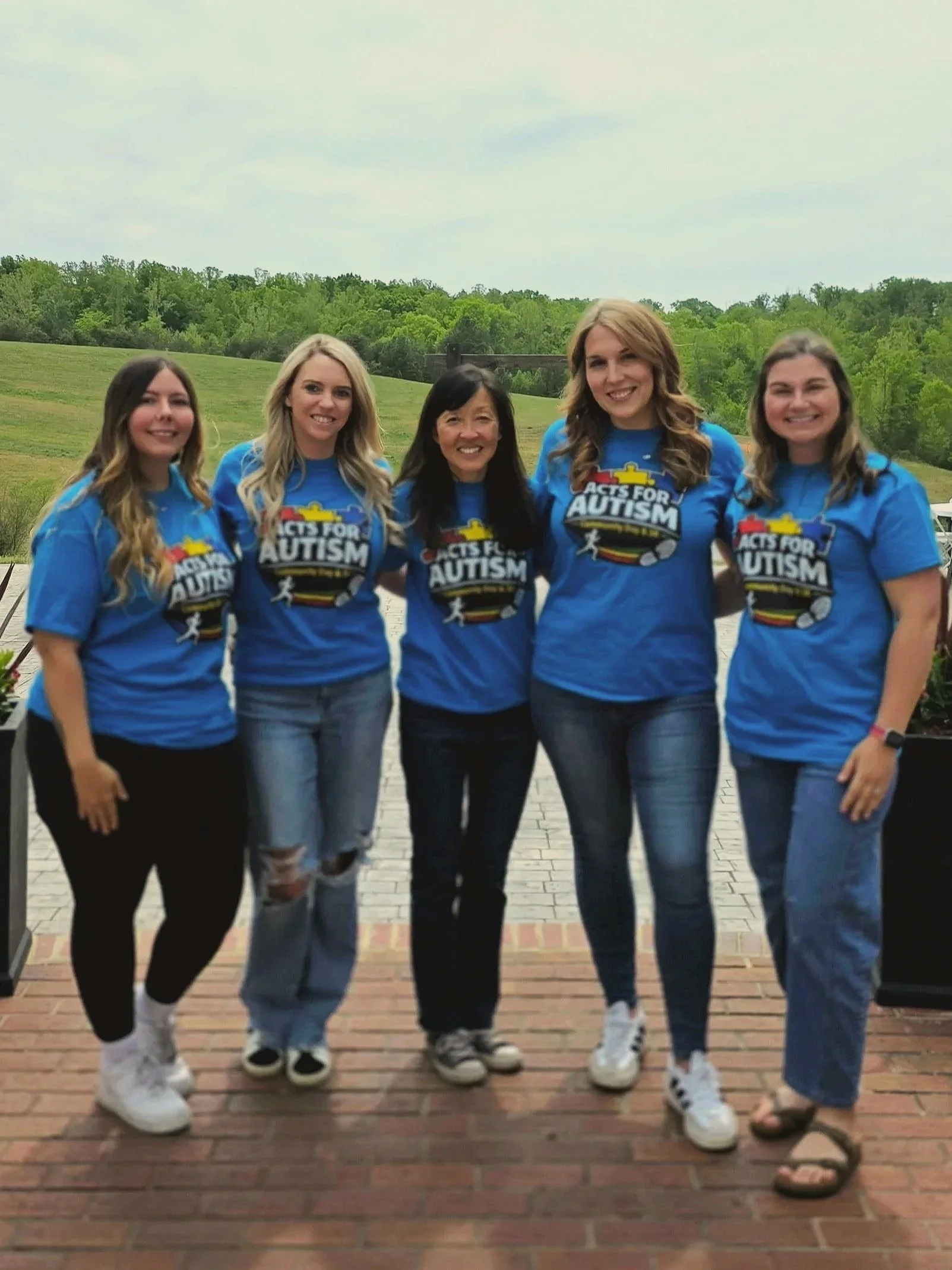 Five women standing together outdoors on a brick patio, wearing blue 'Acts for Autism' shirts, smiling, with green trees and a cloudy sky in the background.