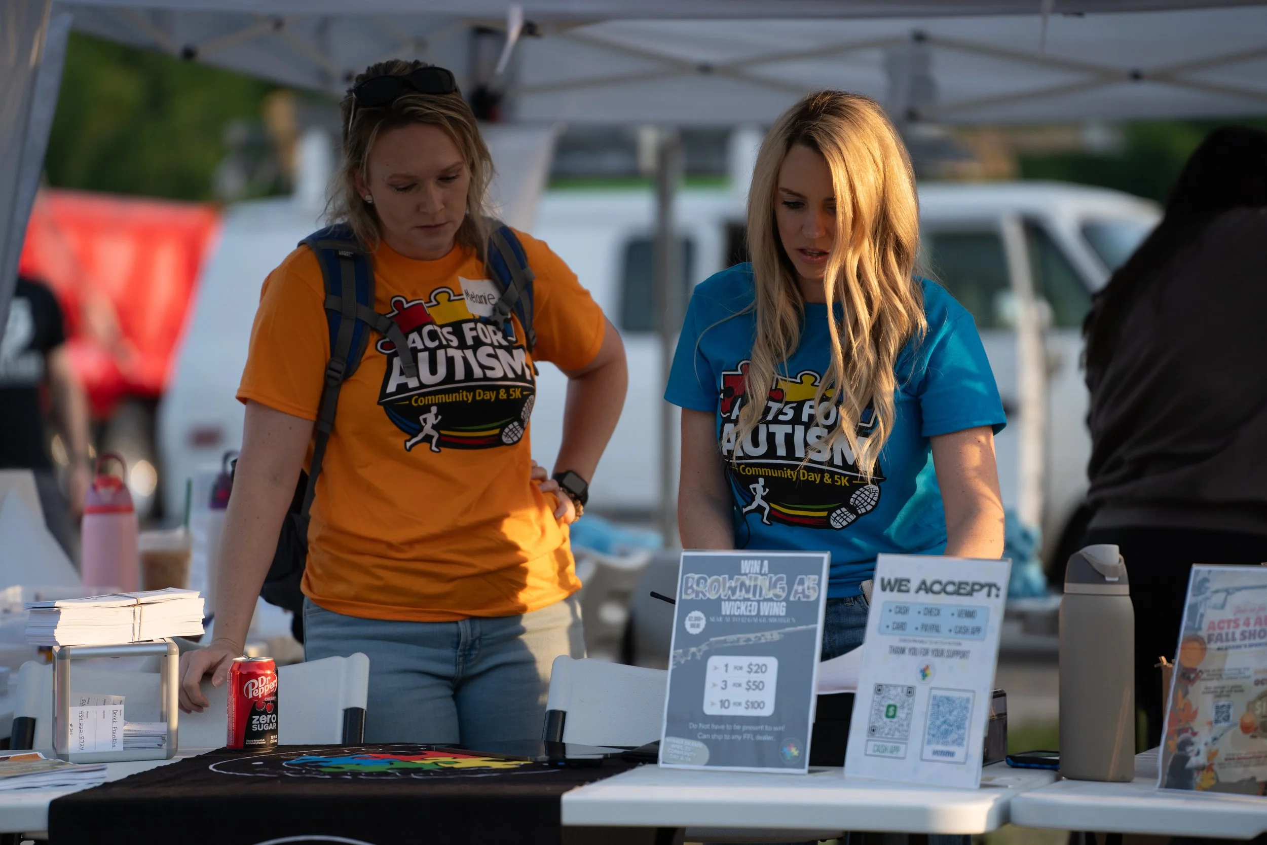 Two women working at a booth during a community event, wearing T-shirts promoting autism awareness, with informational signs and spread-out tables.