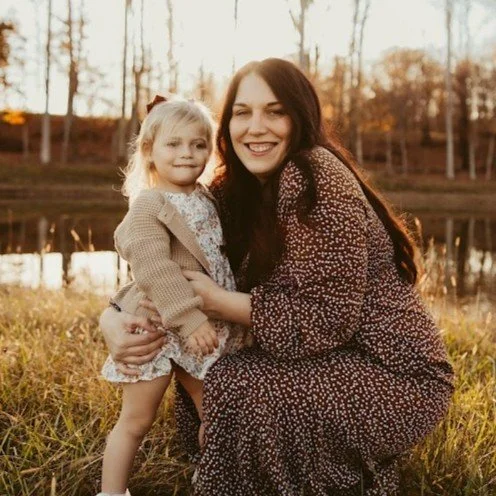 Woman and young girl sitting outdoors near a lake during autumn, smiling at the camera.