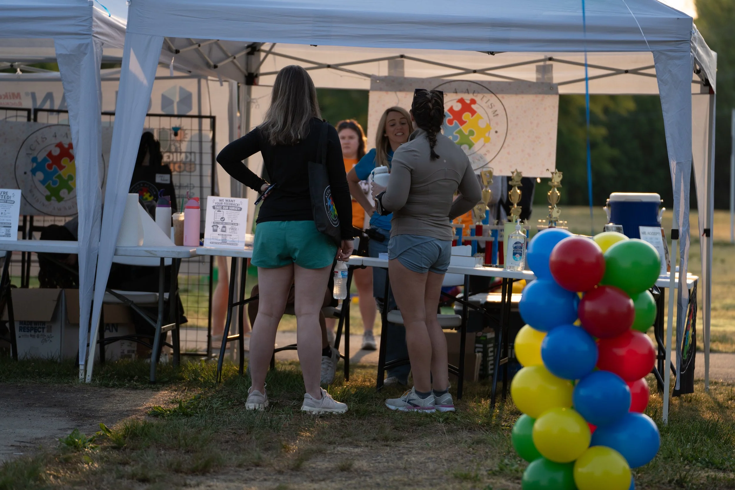 Three women talking at an outdoor event booth decorated with colorful balloons and a banner featuring a multicolored puzzle piece logo.