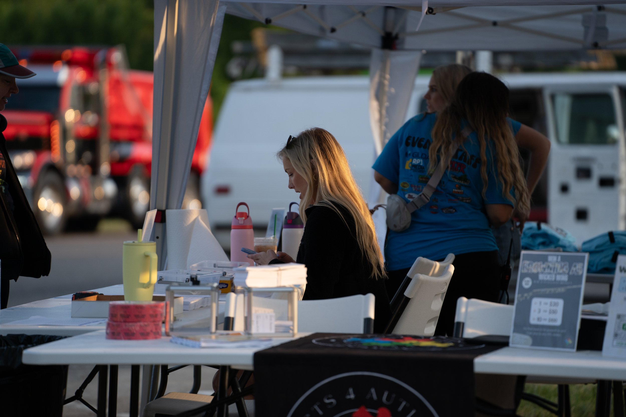 People at an outdoor booth, one person is looking at their phone, the booth has various items and water bottles on the table, with a tent canopy overhead and vehicles in the background.
