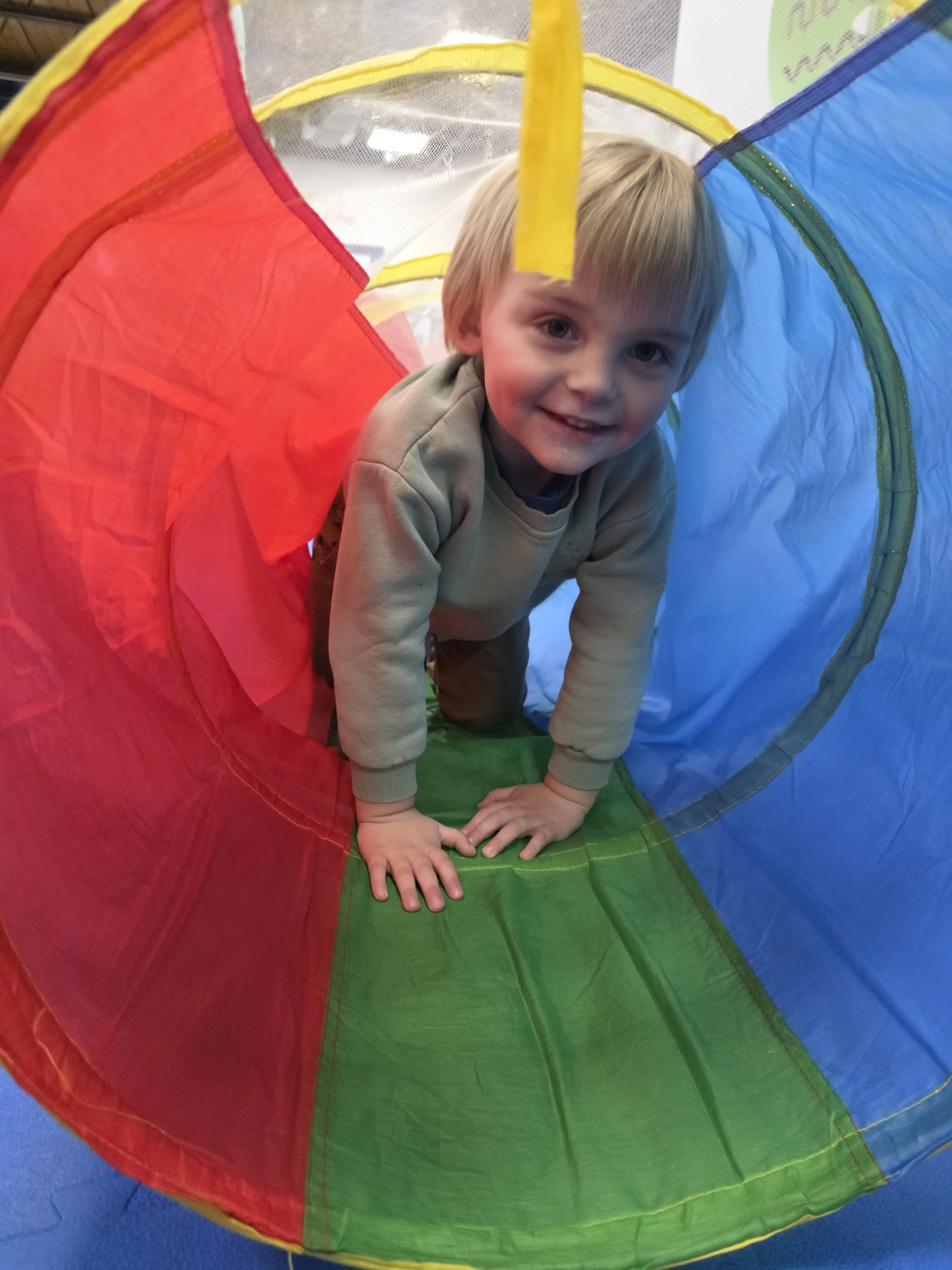 Young boy crawling through a colorful play tunnel with red, green, and blue sections, on a blue floor.