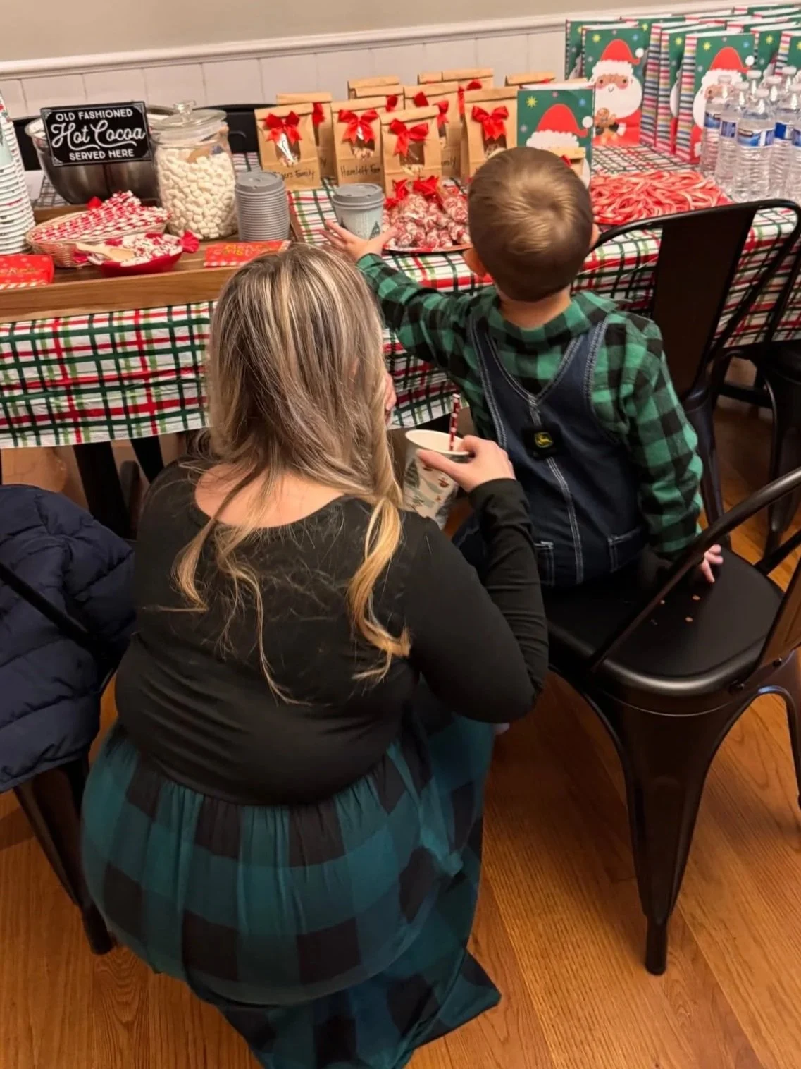 A woman kneeling in front of a young boy at a Christmas party, reaching for the table filled with holiday treats and gifts, decorated with Christmas-themed items.