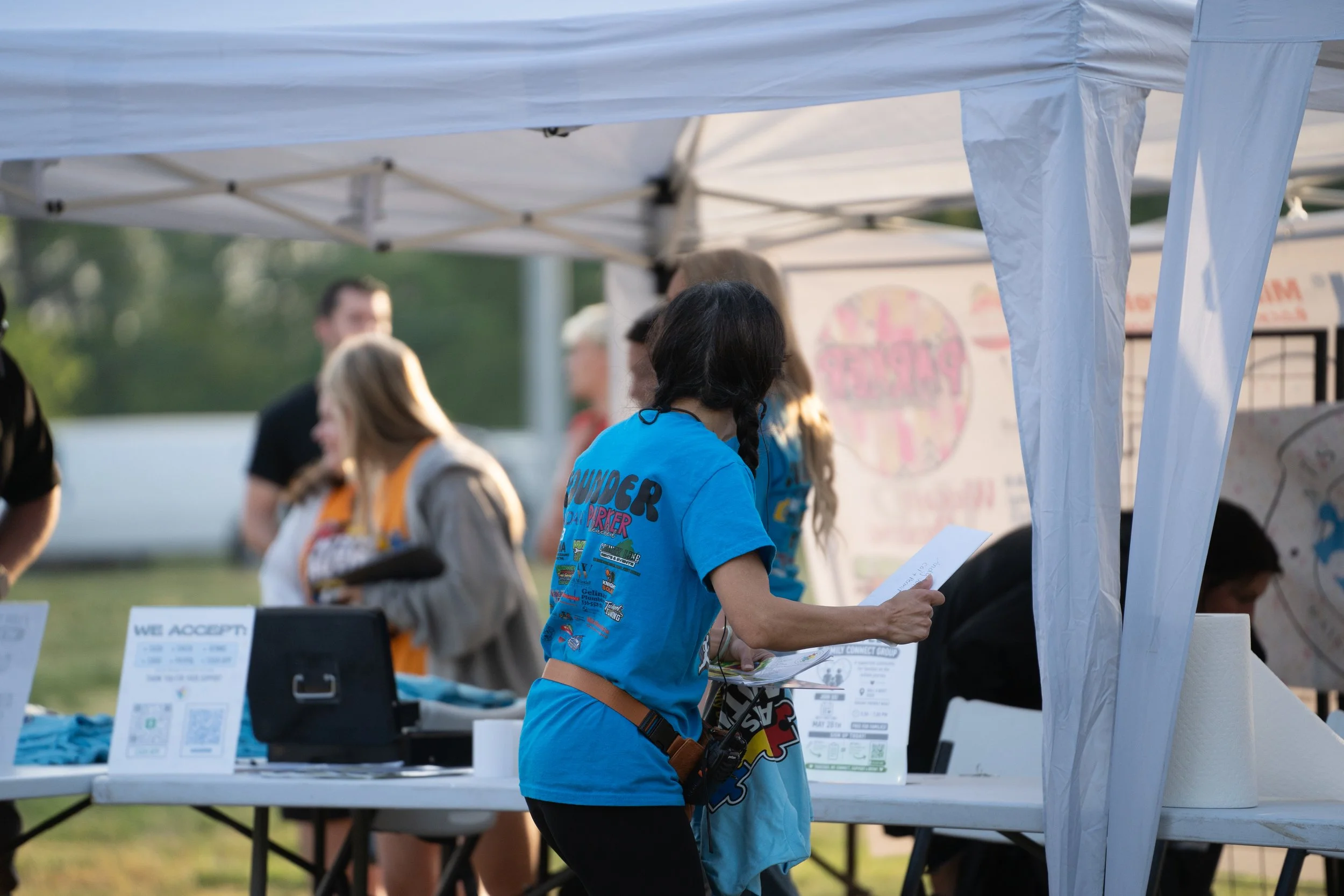 A young girl with braided hair wearing a bright blue t-shirt reading 'Thunder' stands at a table with flyers or papers at an outdoor event under a white tent, with several other people blurred in the background.
