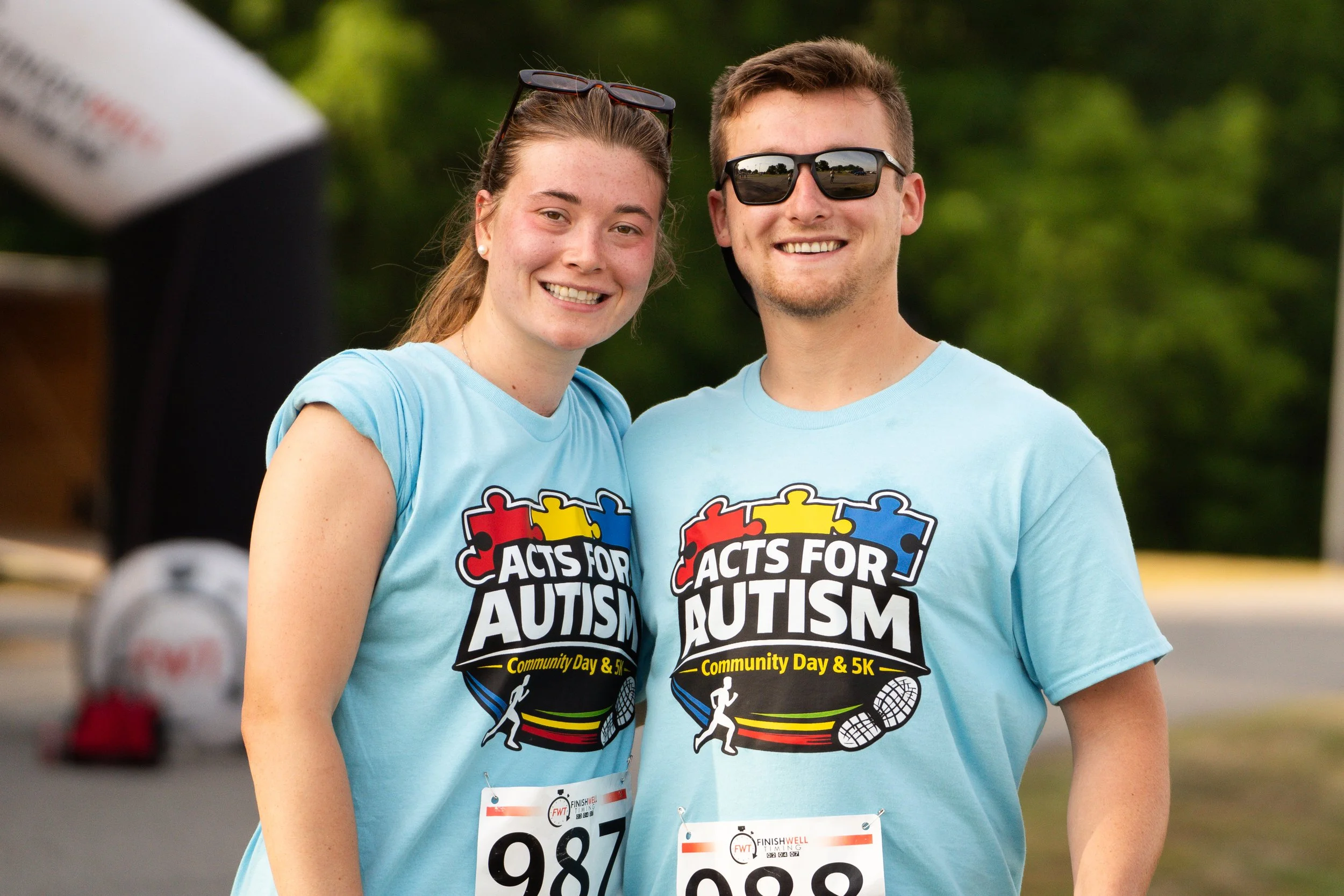 Two smiling young adults, one female and one male, wearing light blue T-shirts with 'Acts for Autism' and event details, standing outdoors at a community event, with green trees and other participants in the background.