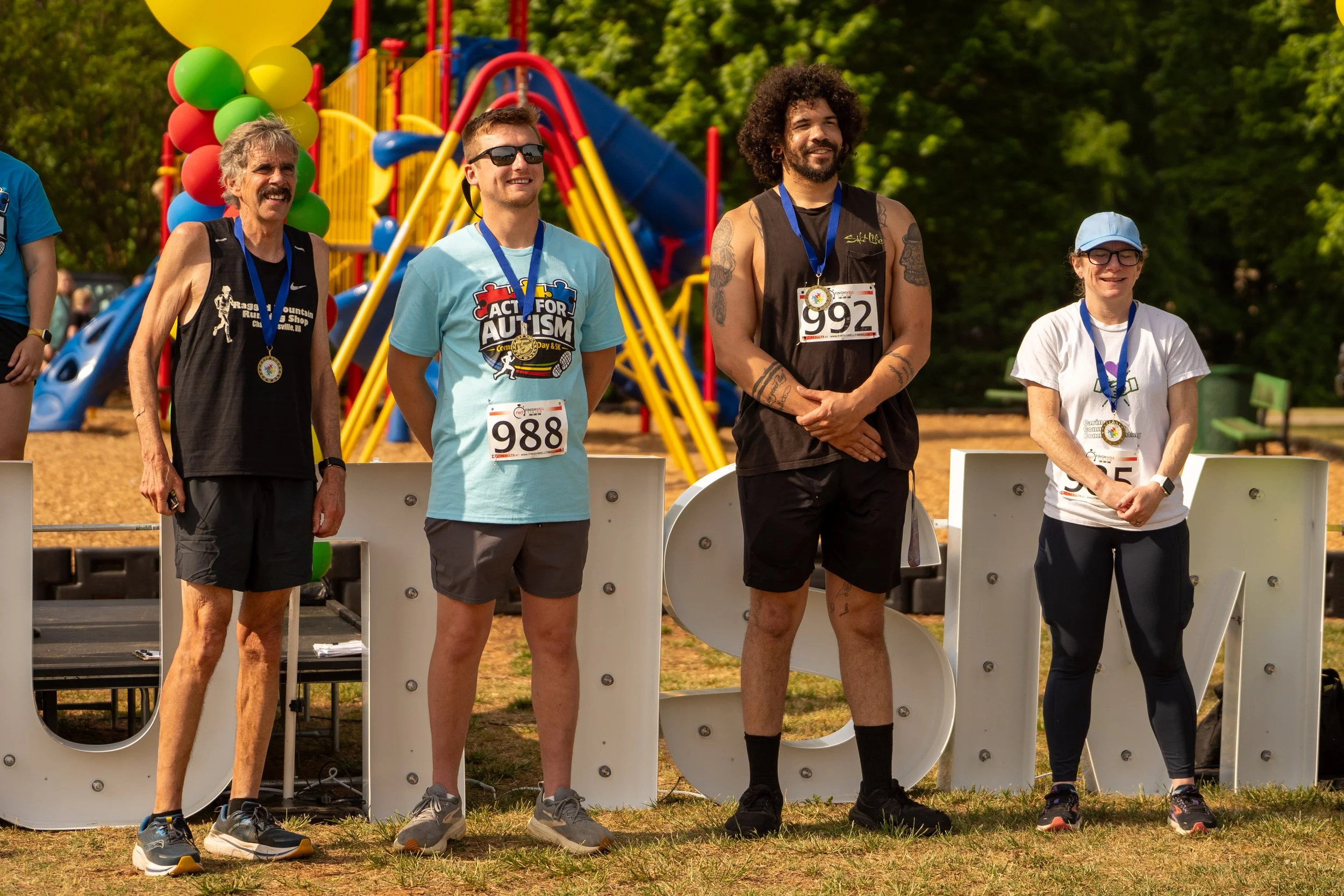 Four individuals standing on an outdoor stage after a race, wearing medals and numbered bibs, with playground equipment and trees in the background.