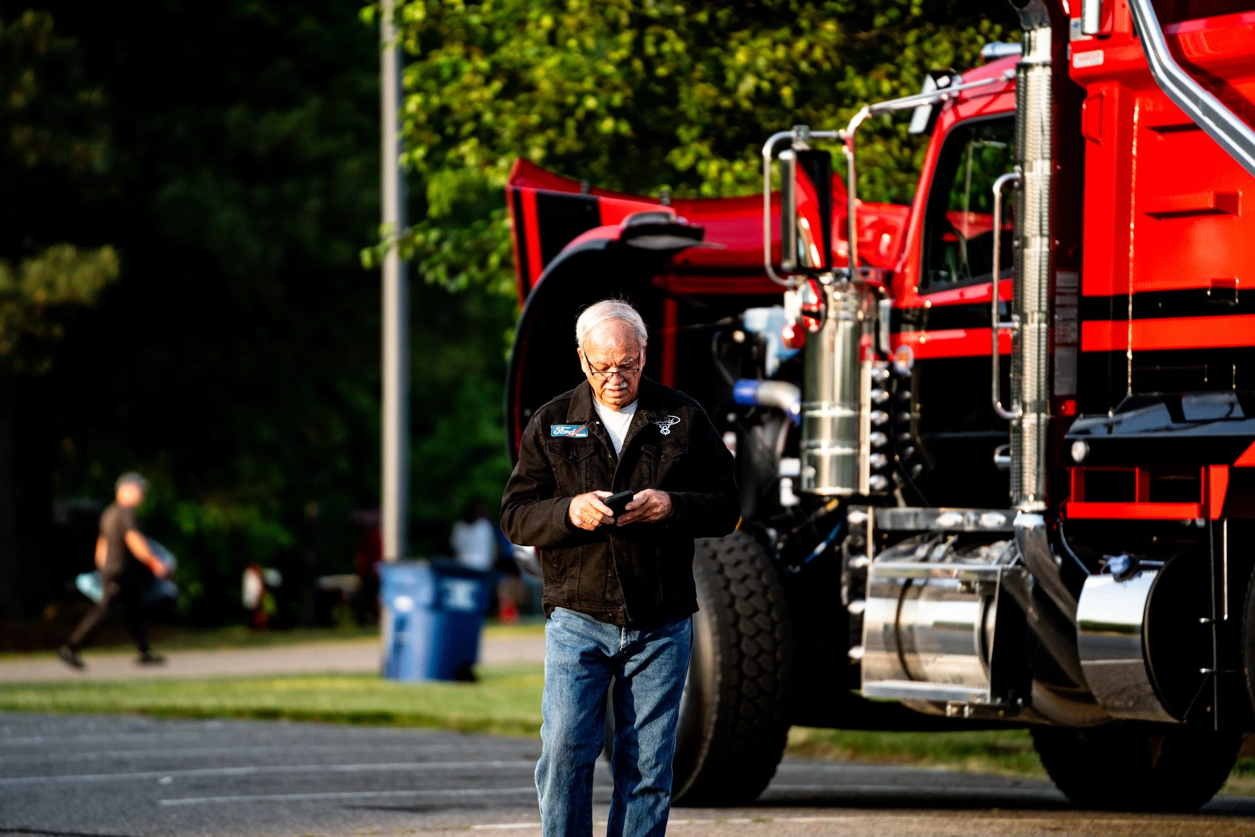 An older man with gray hair and glasses walking past a large red fire truck while looking at his phone in a park or outdoor area.