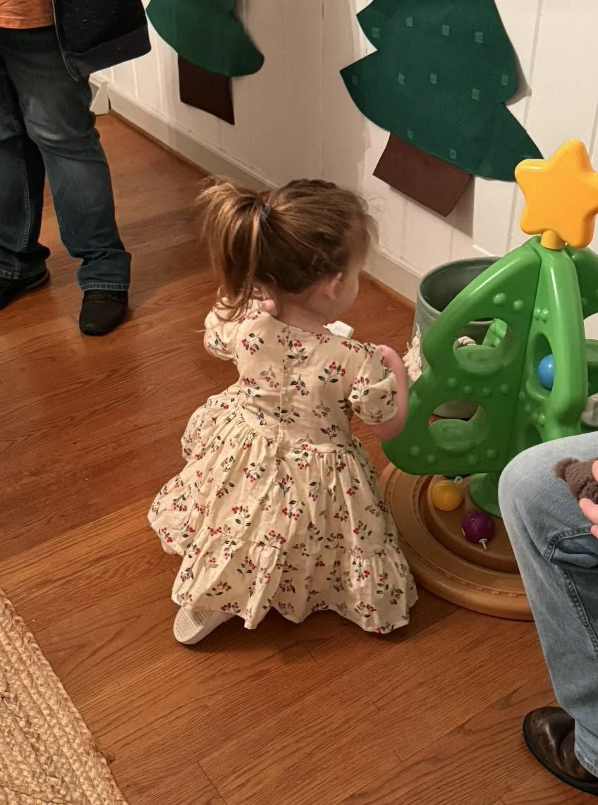 A young girl with a ponytail wearing a floral dress is kneeling on the wooden floor, reaching towards a small green Christmas tree-shaped toy with a yellow star on top, in a room with wooden flooring and paper holiday decorations on the wall.