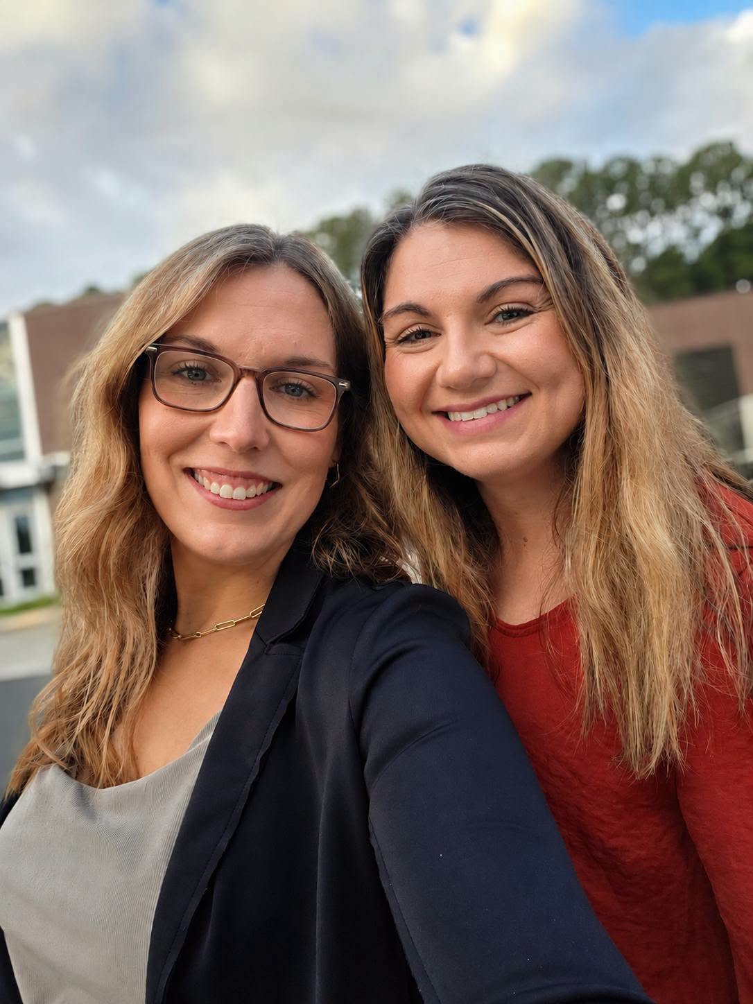 Two smiling women taking a selfie outdoors with trees and buildings in the background.