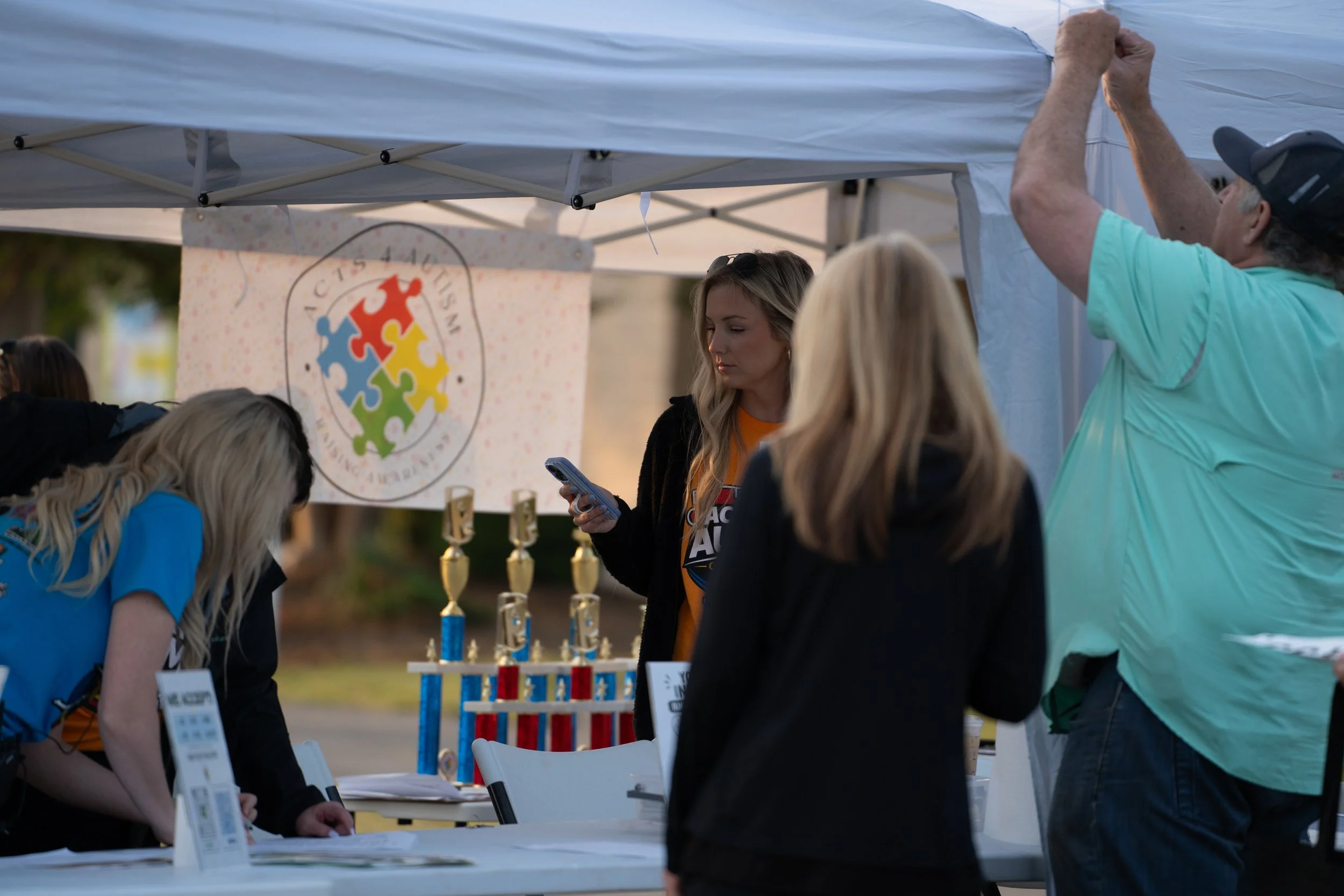 People at an outdoor booth with a large jigsaw puzzle logo, surrounded by tables with awards and informational materials.