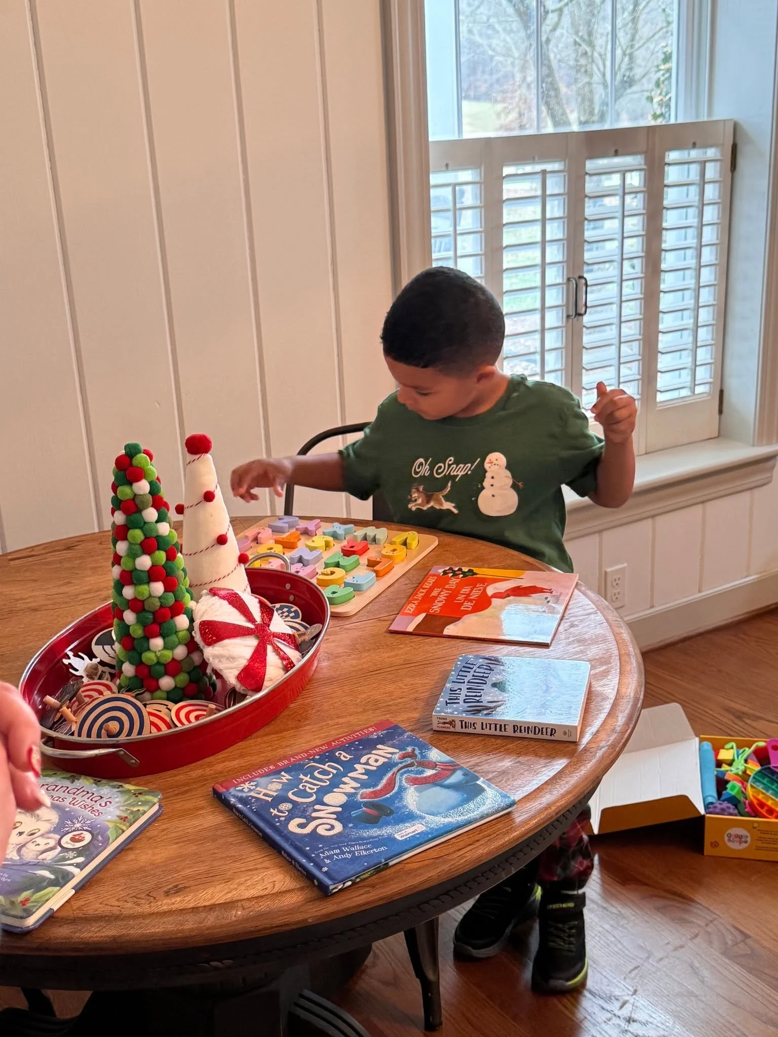 A young boy standing behind a wooden table decorated with Christmas items, including two felt Christmas trees, holiday books, and toys, with a window in the background.