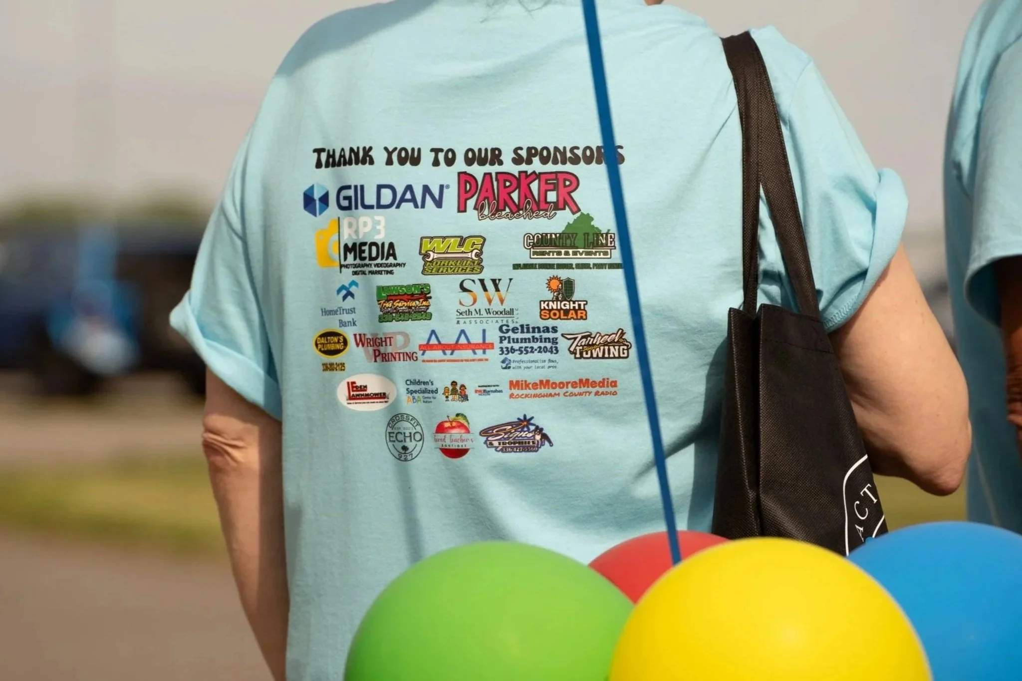 Back of a person wearing a light blue T-shirt with various sponsor logos, standing outdoors near colorful balloons.
