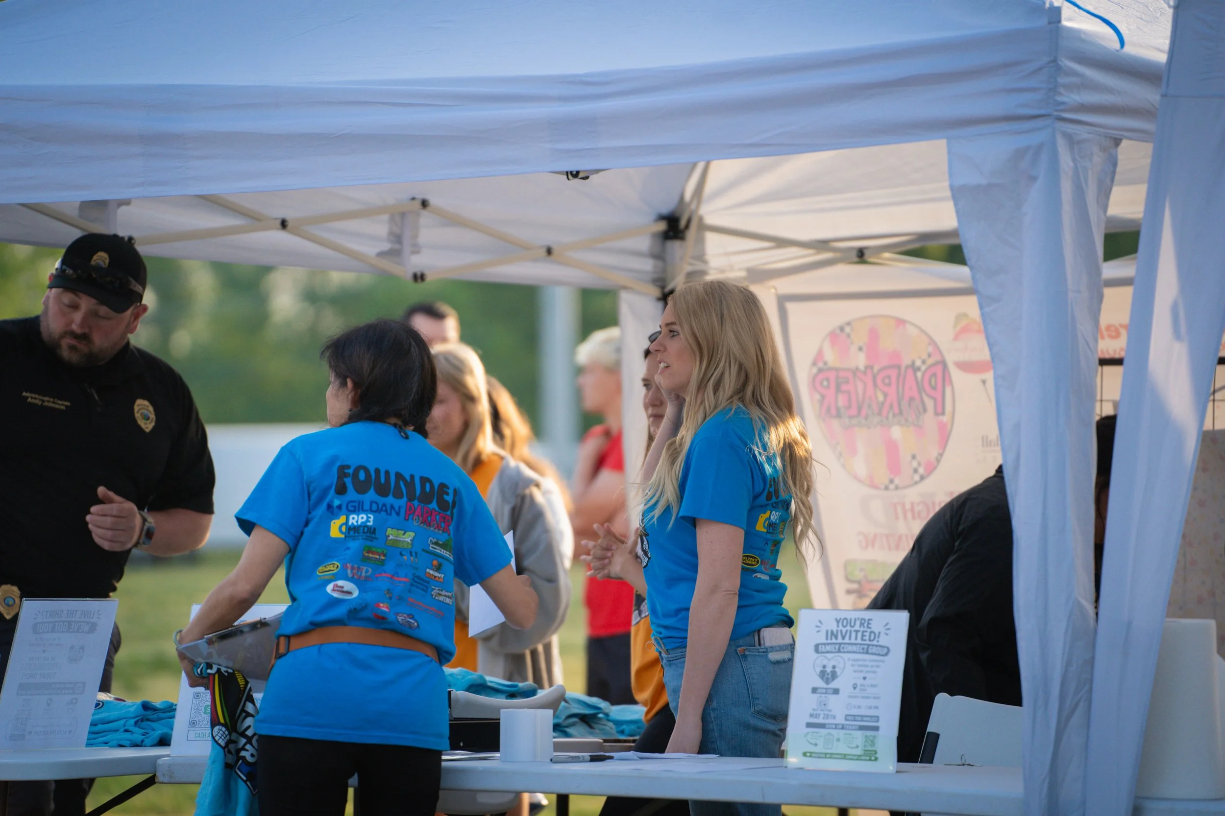 Group of people under a white canopy tent at an outdoor event, some wearing blue shirts with sponsor logos, engaging in conversation, with informational signs and tables in front.