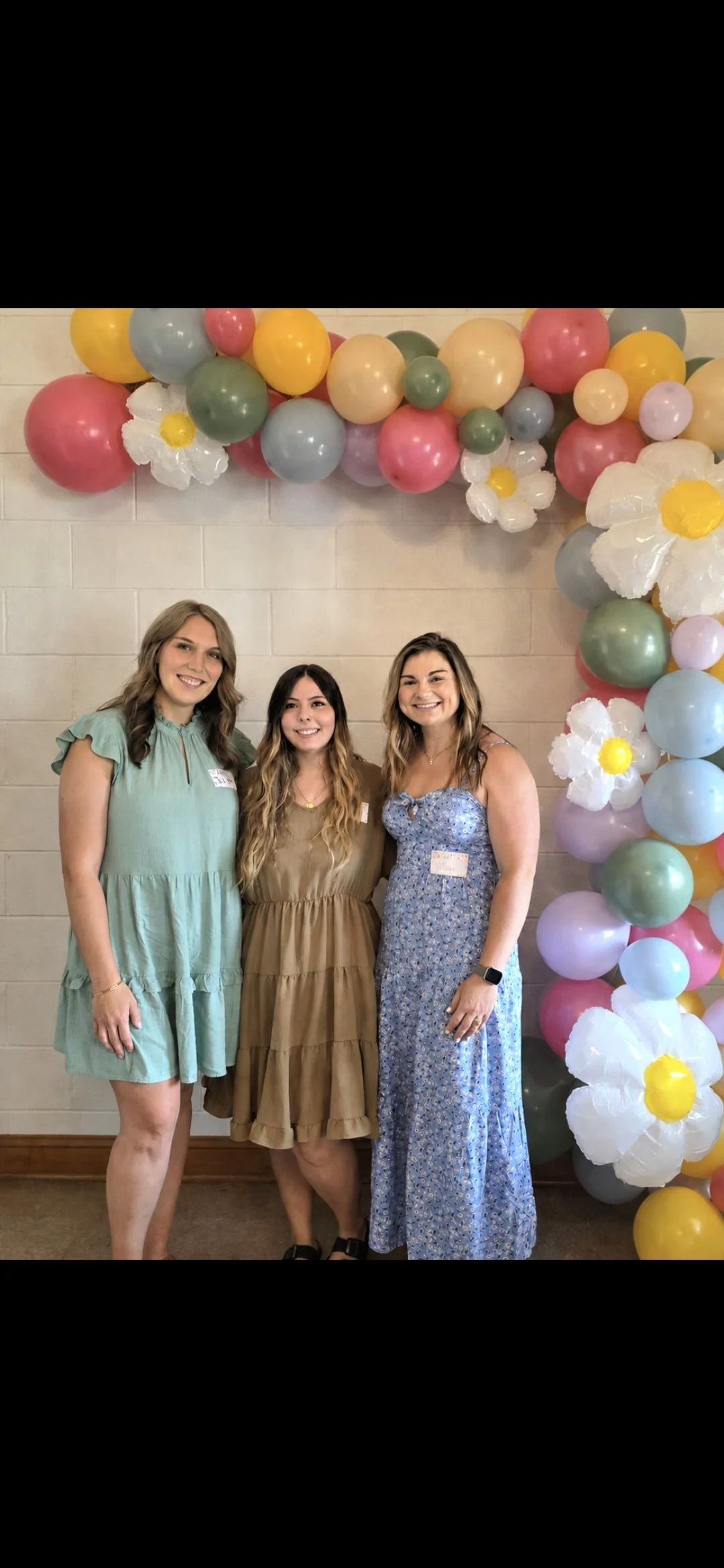 Three women standing together smiling in front of a colorful balloon arch with flowers, beige wall, and wooden floor.