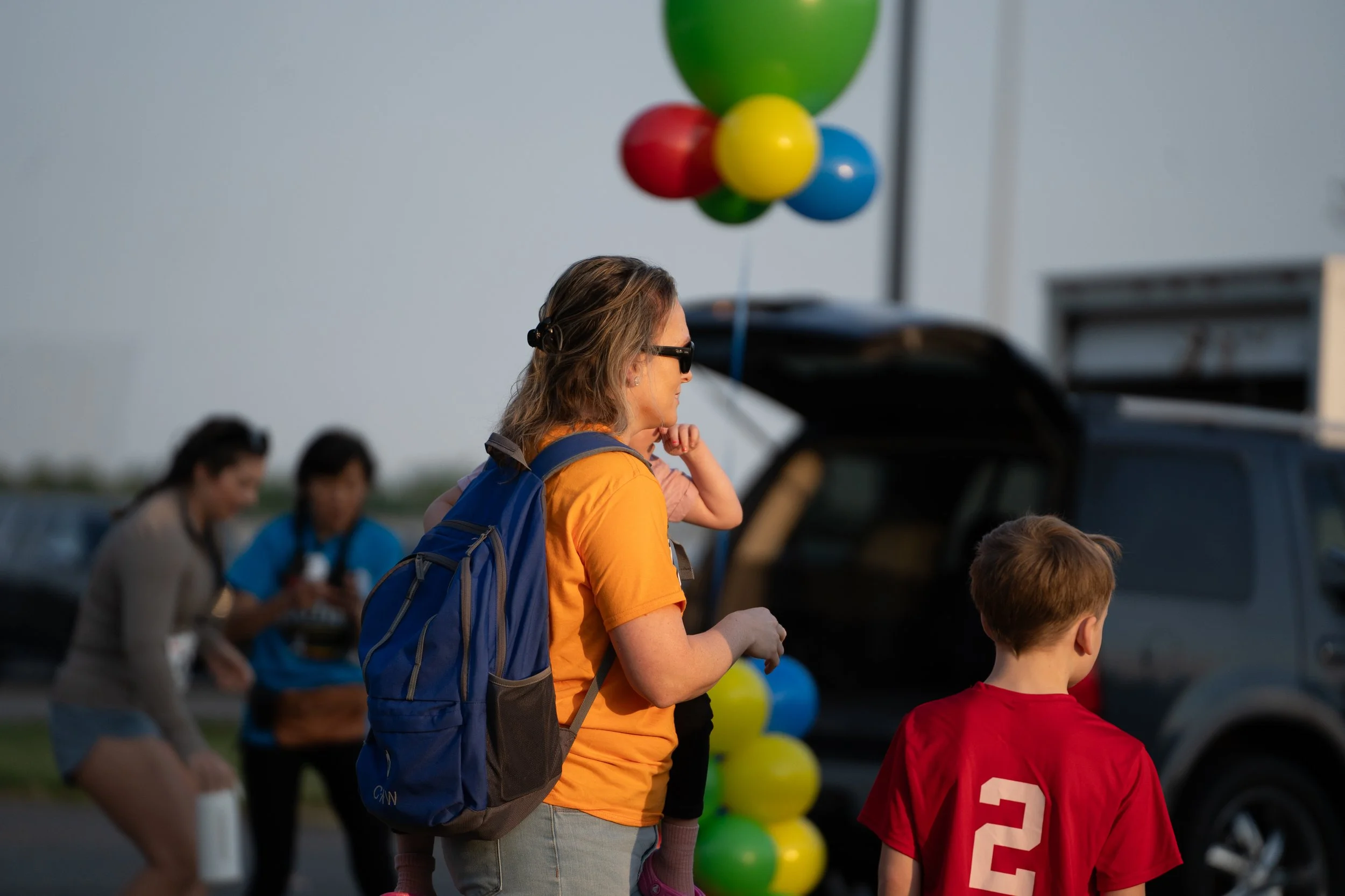 A woman with sunglasses, wearing an orange shirt and carrying a blue backpack, stands next to a boy wearing a red sports jersey, at an outdoor event with balloons in the background.