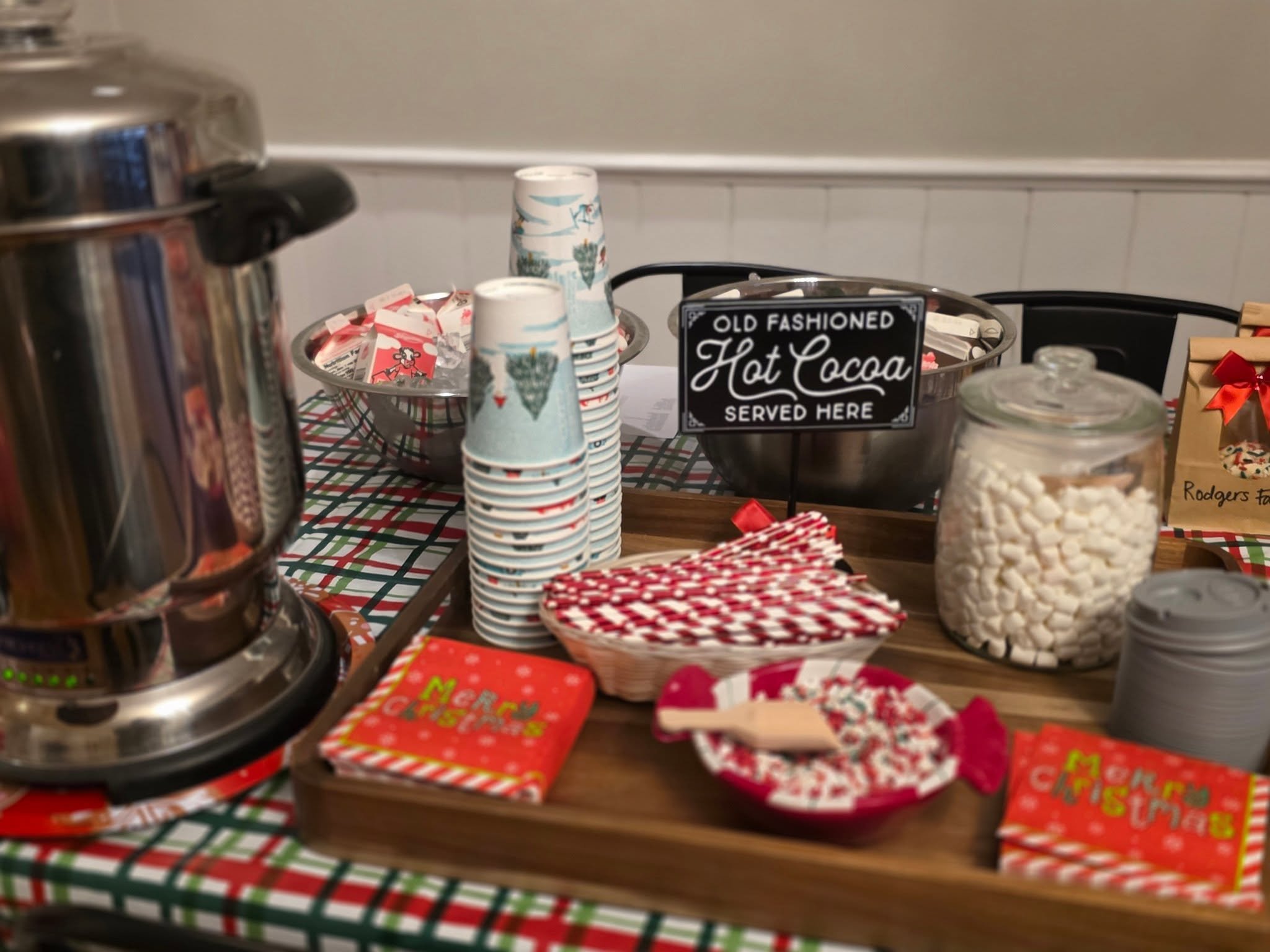 A table with Christmas-themed items including cups, a sign that reads 'Old Fashioned Hot Cocoa Served Here,' a jar of white marshmallows, wrapped treats, and red and green decorations.