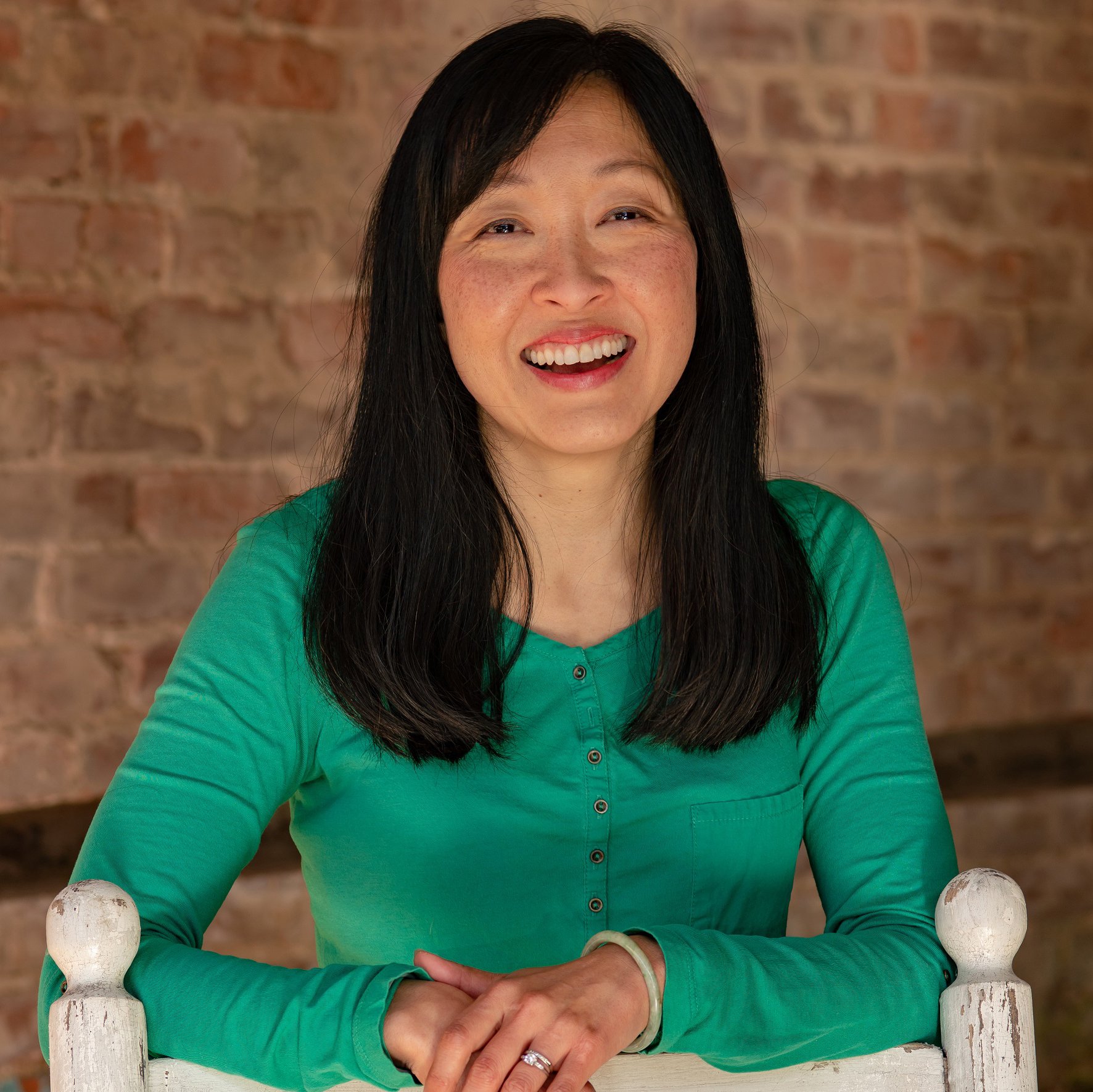 Woman with long black hair in a green shirt sitting on a white wooden bench in front of a brick wall, smiling and looking at the camera.