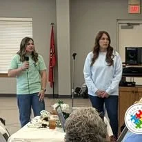 Two women standing at the front of a room, one holding a microphone, in front of seated audience members, with a red flag and a window in the background.