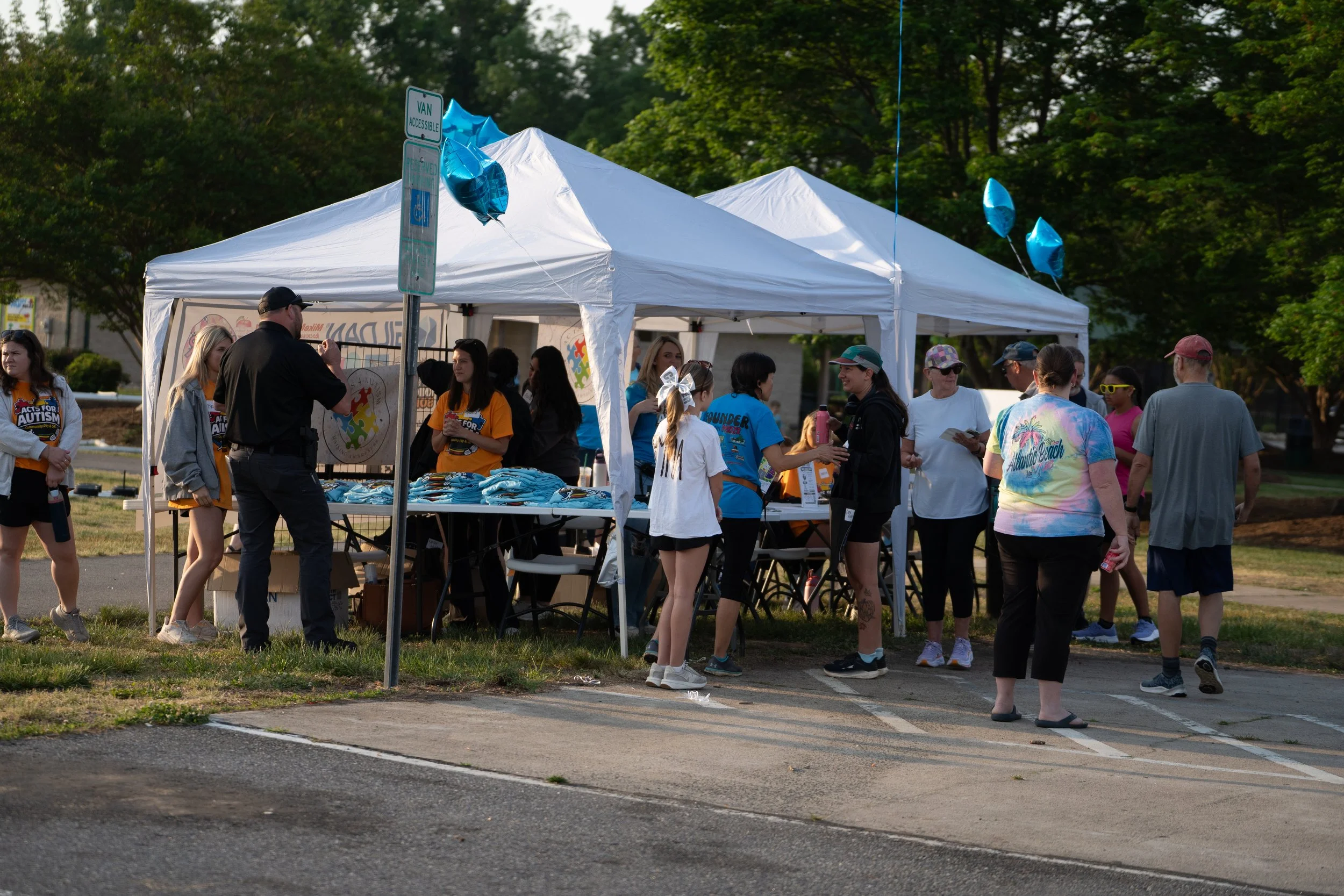 A group of people gathered around a white canopy tent outdoors, some wearing bright event shirts, with balloons attached to the tent and trees in the background.