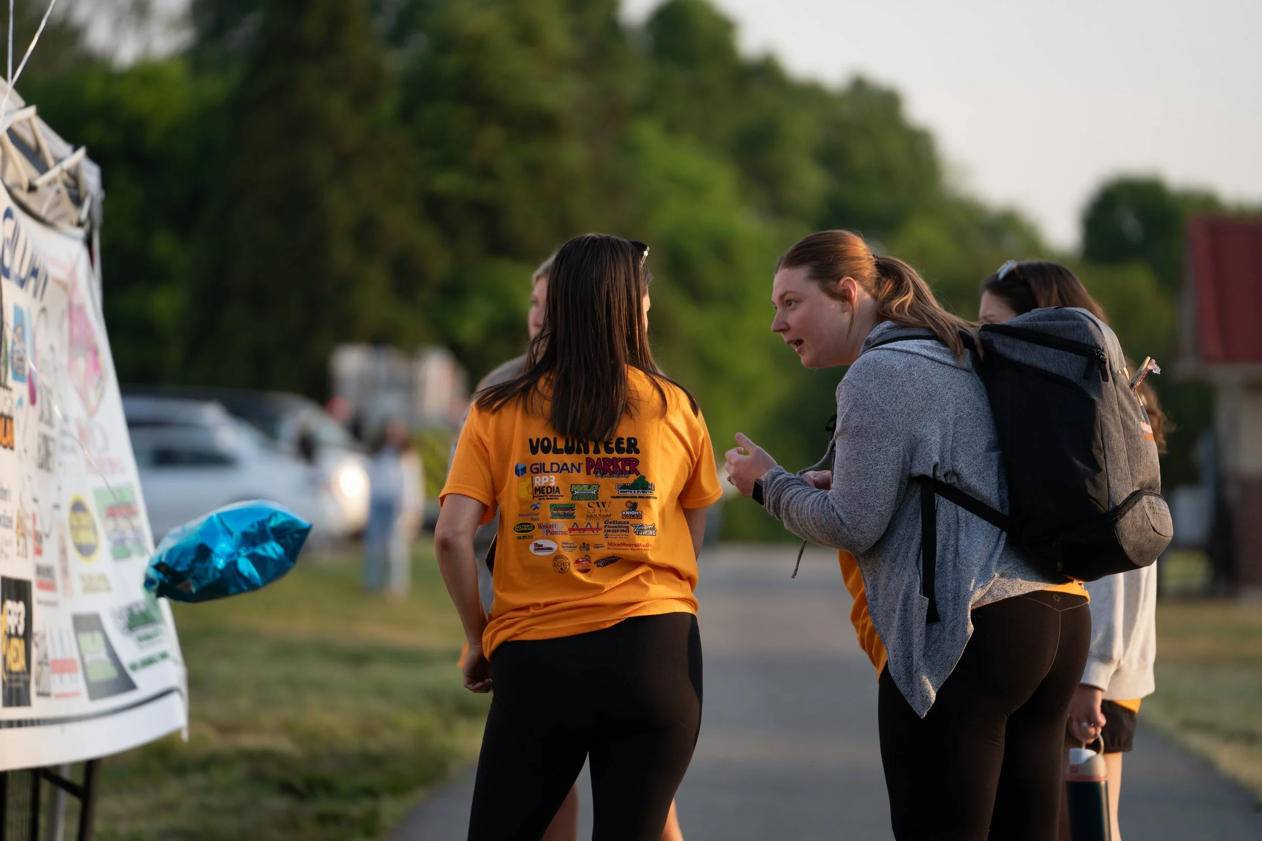 Two women having a conversation outdoors during a community event, with one wearing a volunteer shirt, near a table with stickers and posters.