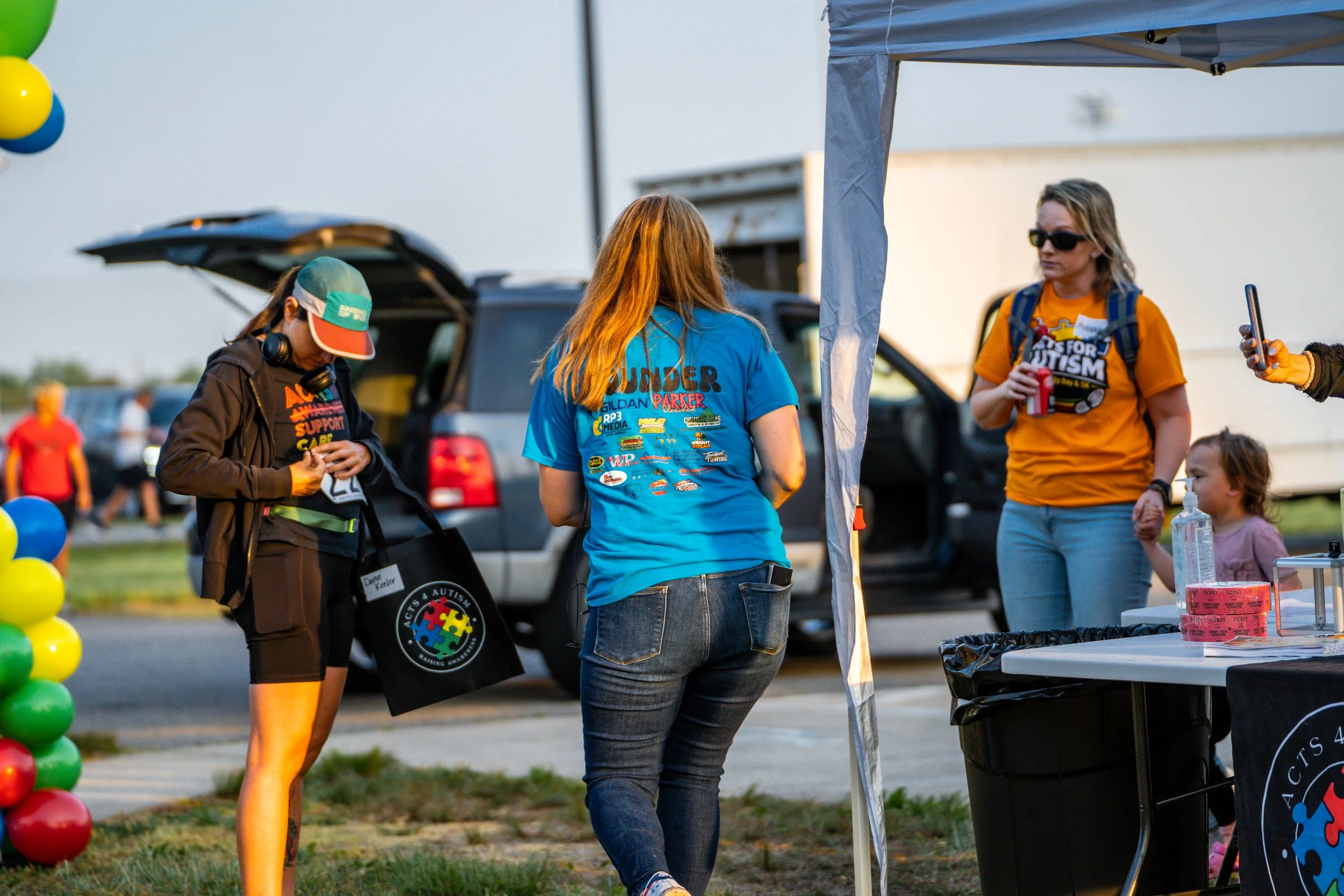 People gathered at an outdoor event, with a man and woman near an autism awareness booth. The booth displays pink tickets and a large black container, and balloons are visible on the left side. In the background, there's a car and other attendees.