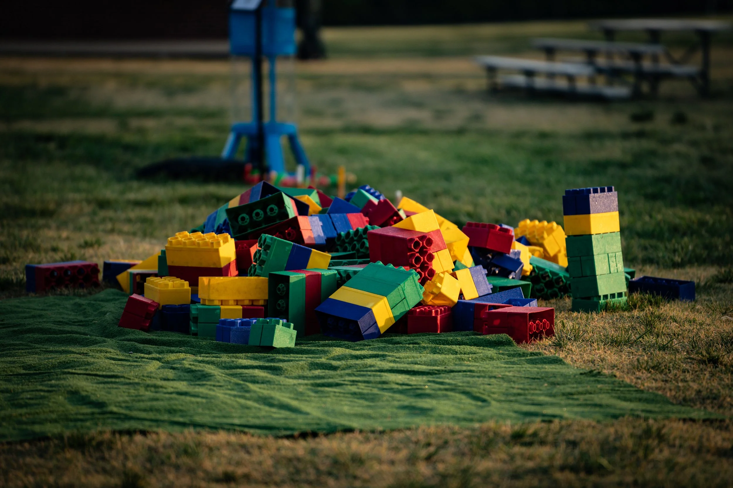 Colorful plastic building blocks scattered on a grassy area at dusk, with empty benches and a blue sculpture in the background.