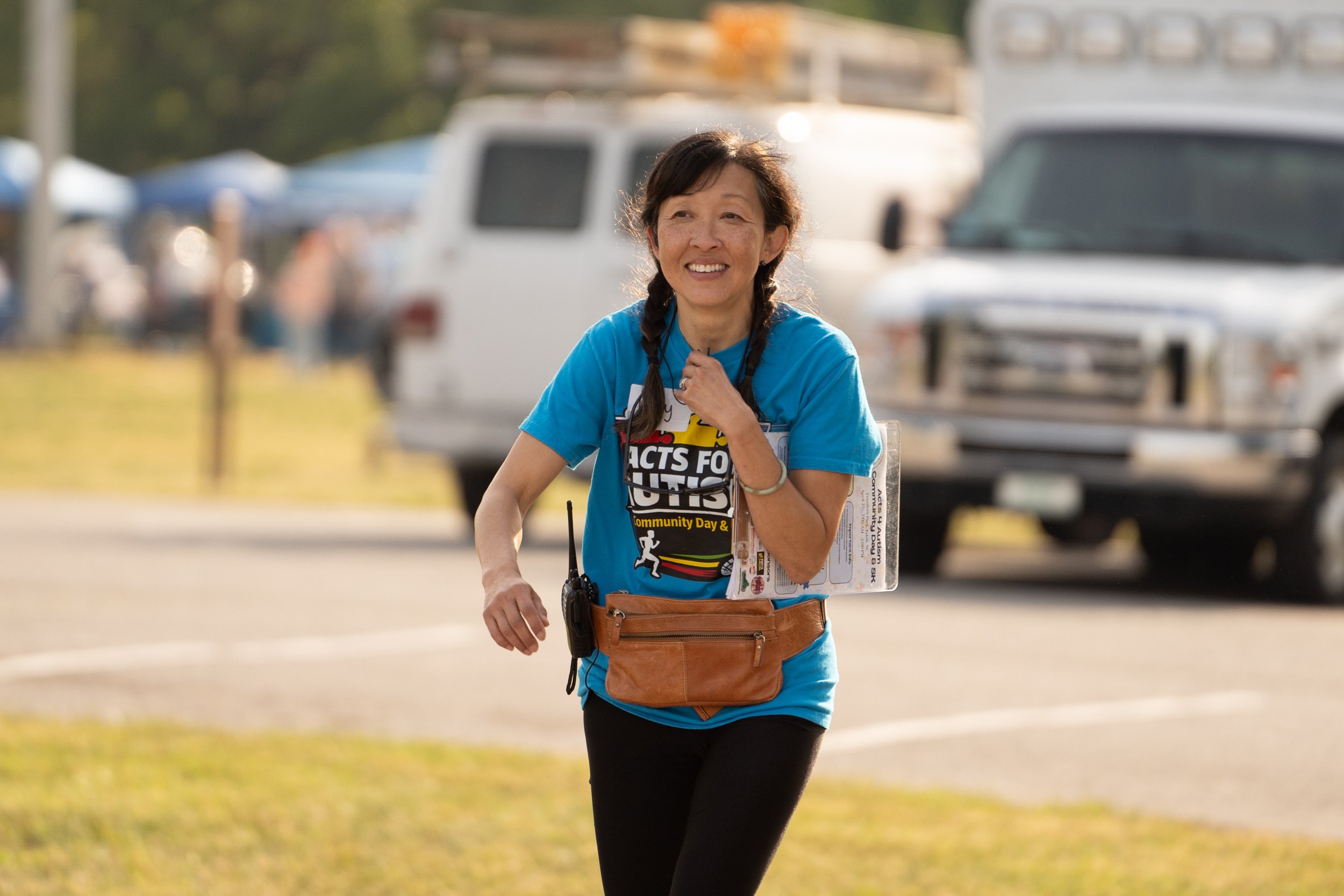 A woman smiling while running outdoors during a community event in the daytime. She is wearing a blue shirt with event text, a brown waist pack, and has a clipboard or event pamphlet in her hand. Background includes vehicles and a loosely focused outdoor setting.