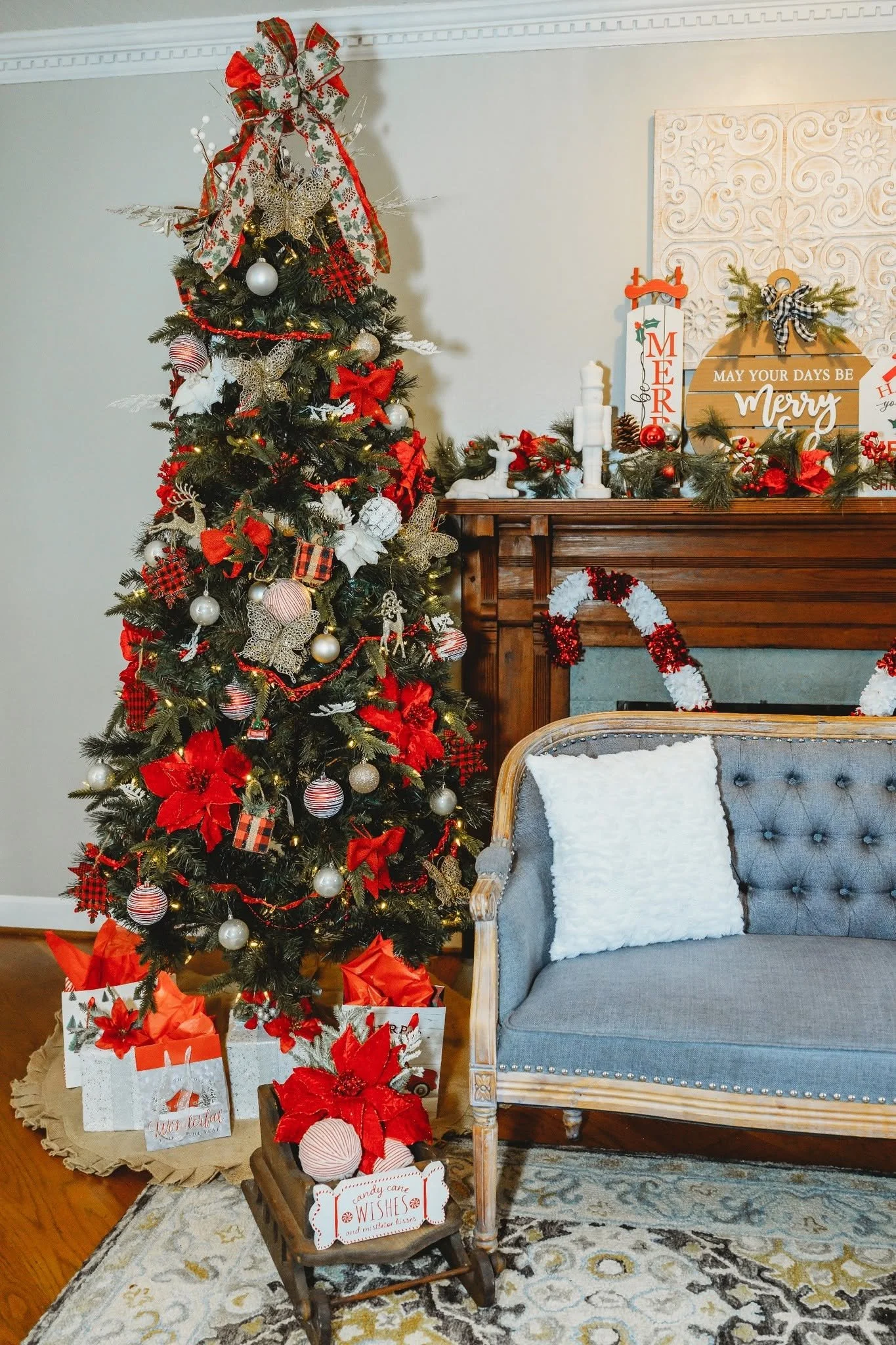 A decorated Christmas tree with ornaments and ribbons next to a wooden fireplace mantel with holiday decor, a blue velvet armchair with a white pillow, and holiday signs and gift bags.