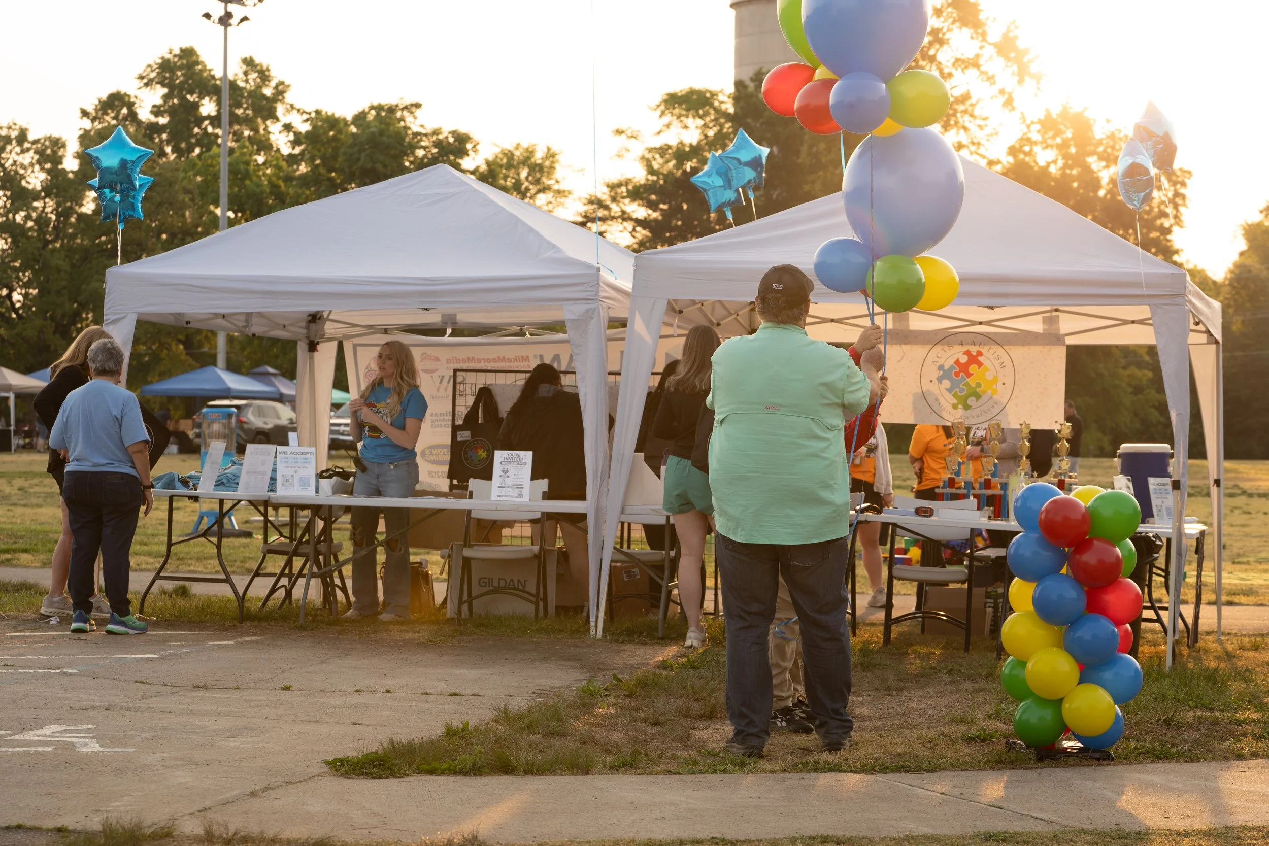 People at an outdoor event with white tents decorated with colorful balloons, some people are talking, and the sun is setting in the background.