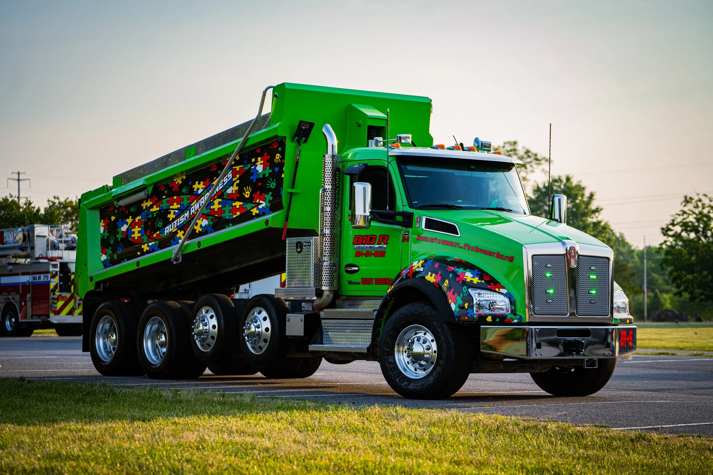 A bright green truck with autism puzzle piece graphics, parked outdoors with trees and a clear sky in the background.