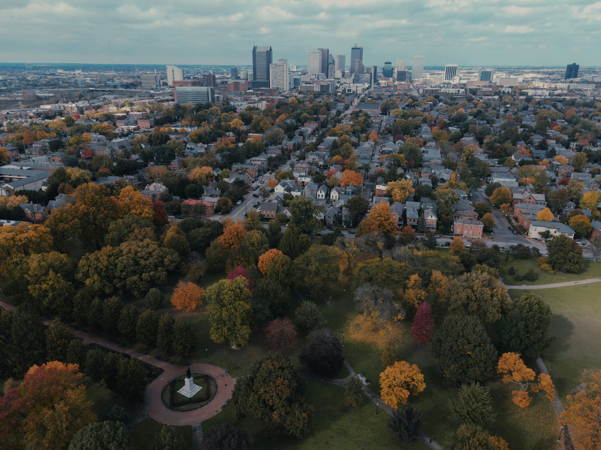 Aerial view of the Cincinnati skyline with tall buildings in the background and a residential neighborhood with autumn-colored trees in the foreground. There is a park with a monument at the bottom of the image.