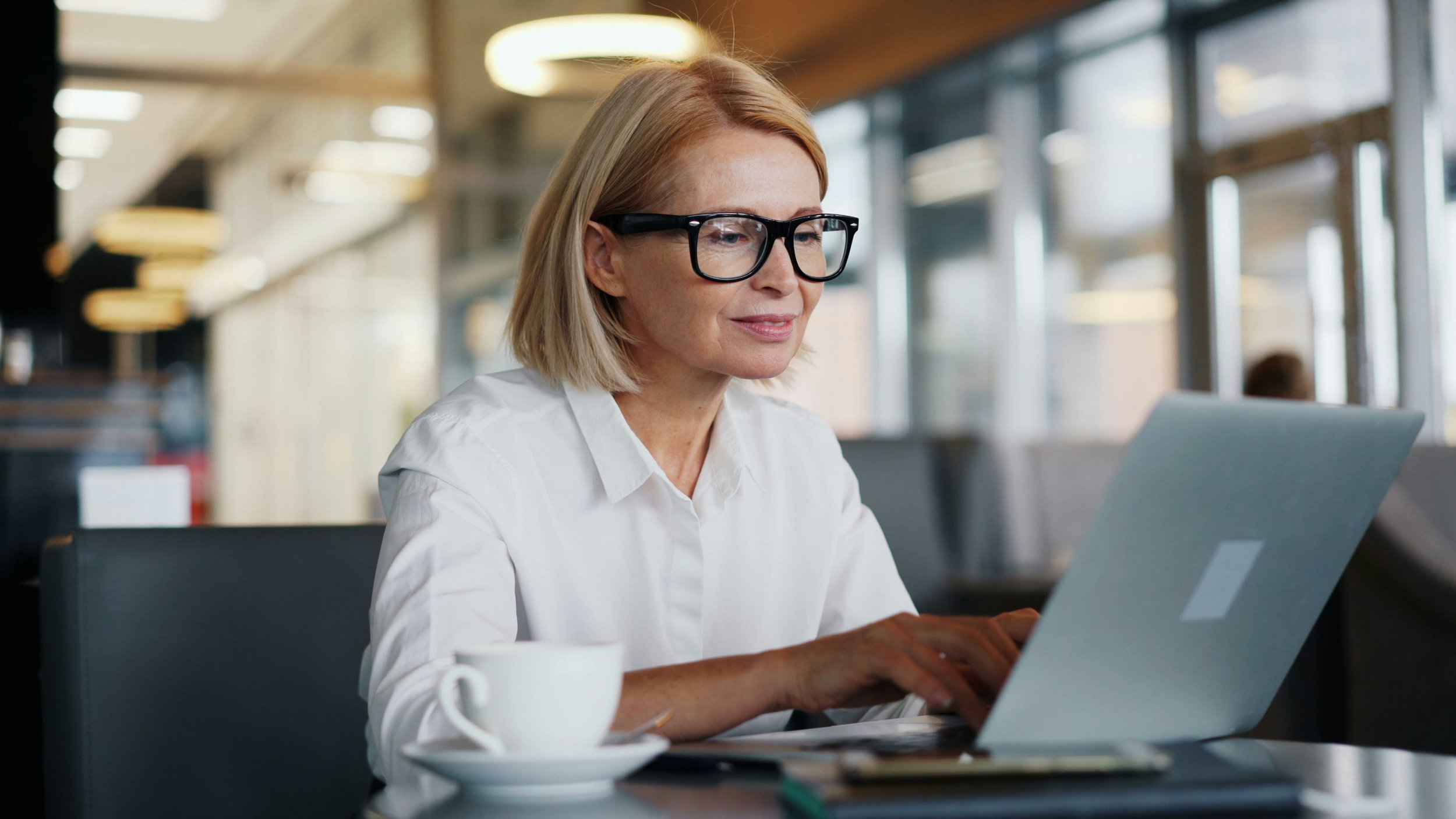 A woman with blonde hair, wearing large black glasses and a white shirt, working on a laptop in a modern, well-lit office or café.