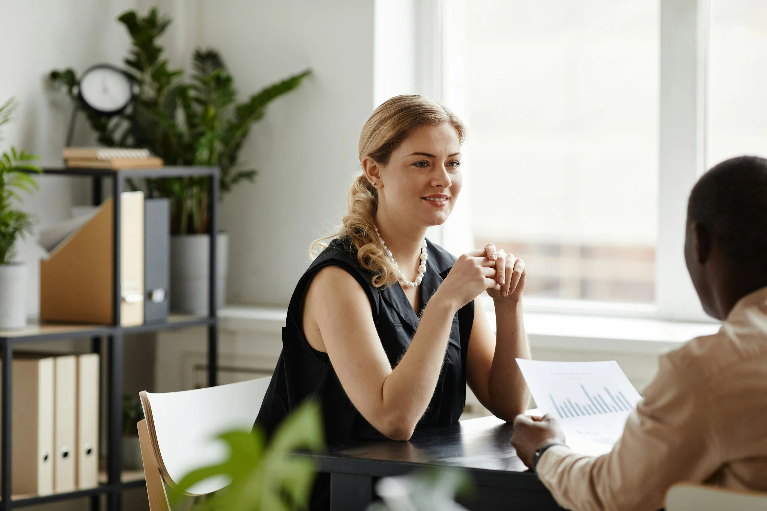 A woman with blonde hair, wearing a black top and pearl necklace, smiling and sitting across from a person in a beige shirt holding a document, in a modern office setting with plants and a clock in the background.