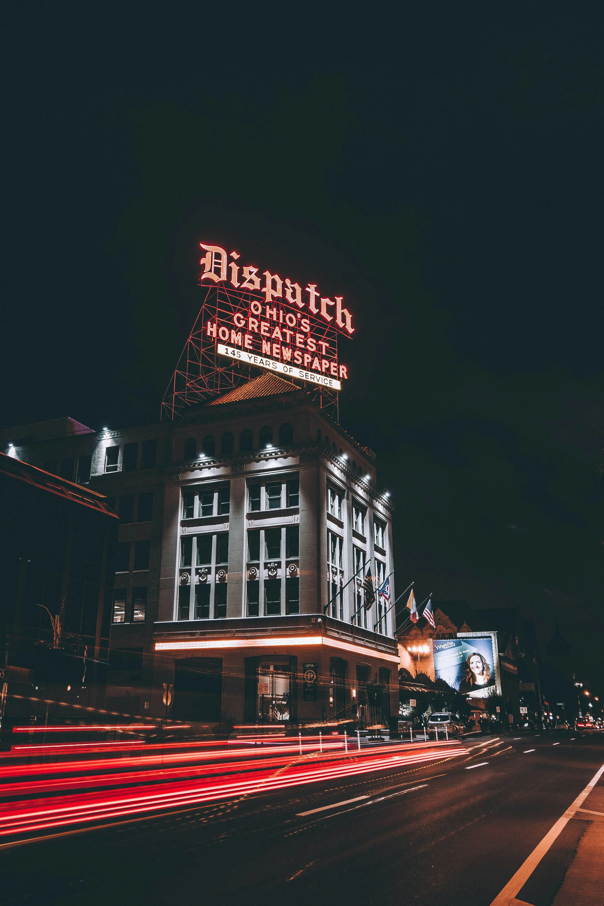 Illuminated sign above a building on a Cincinnati street at night, with light trails from passing traffic