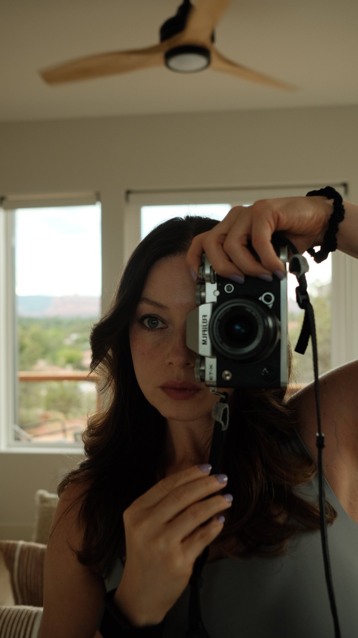 A woman with dark hair taking a mirror selfie with a camera, with view of windows and landscape in the background.