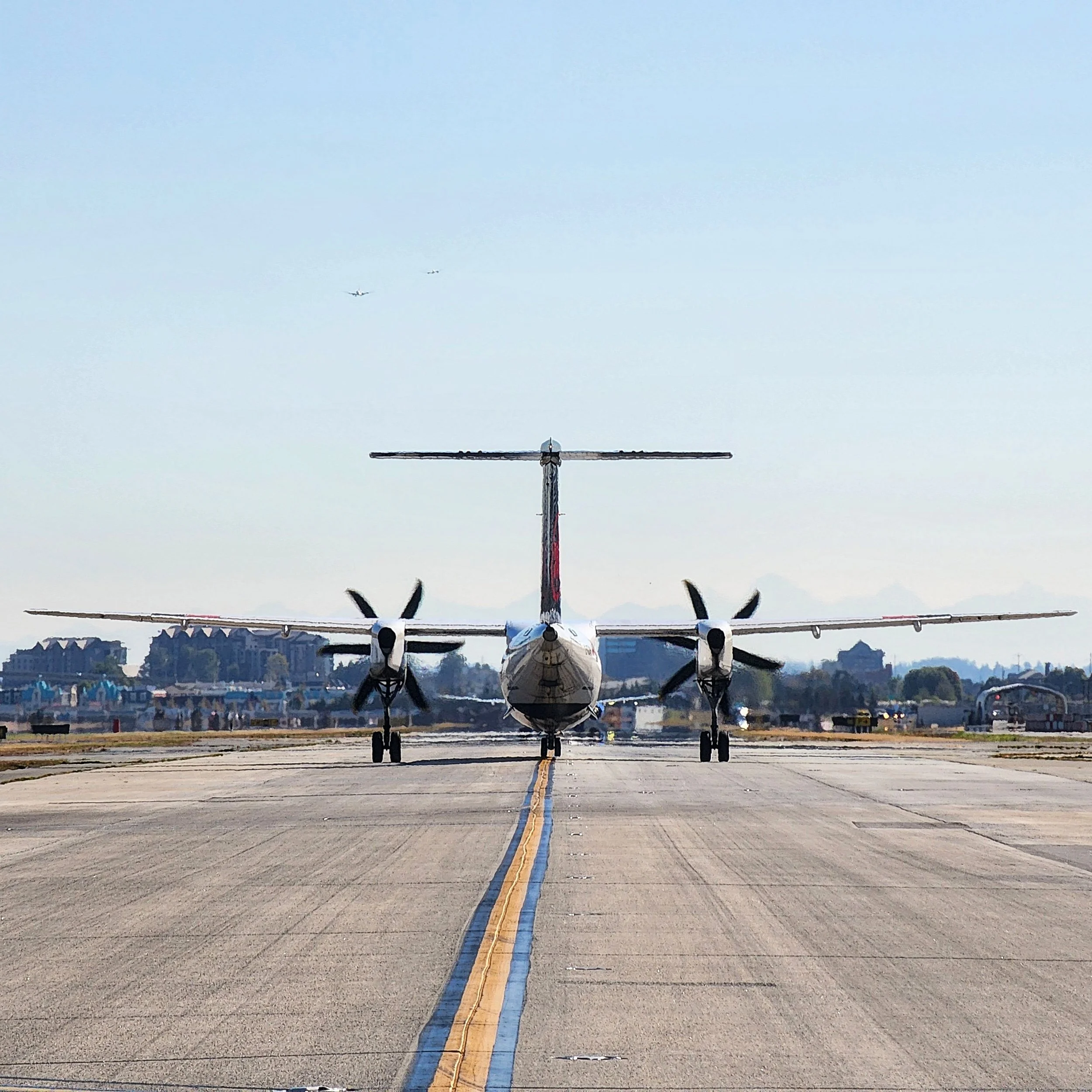 An airplane is on a runway with two smaller planes flying overhead. The runway has a yellow and blue center line, and there are mountains and trees in the background.