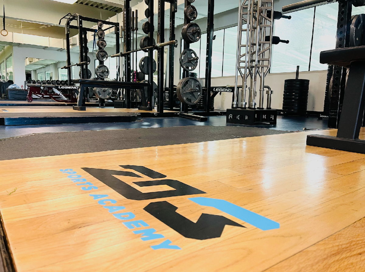 Empty gym with weightlifting equipment, black weight plates, barbells, and a wooden platform with the ACXDEMY SPORTS logo in blue and black, large windows in the background letting in natural light.