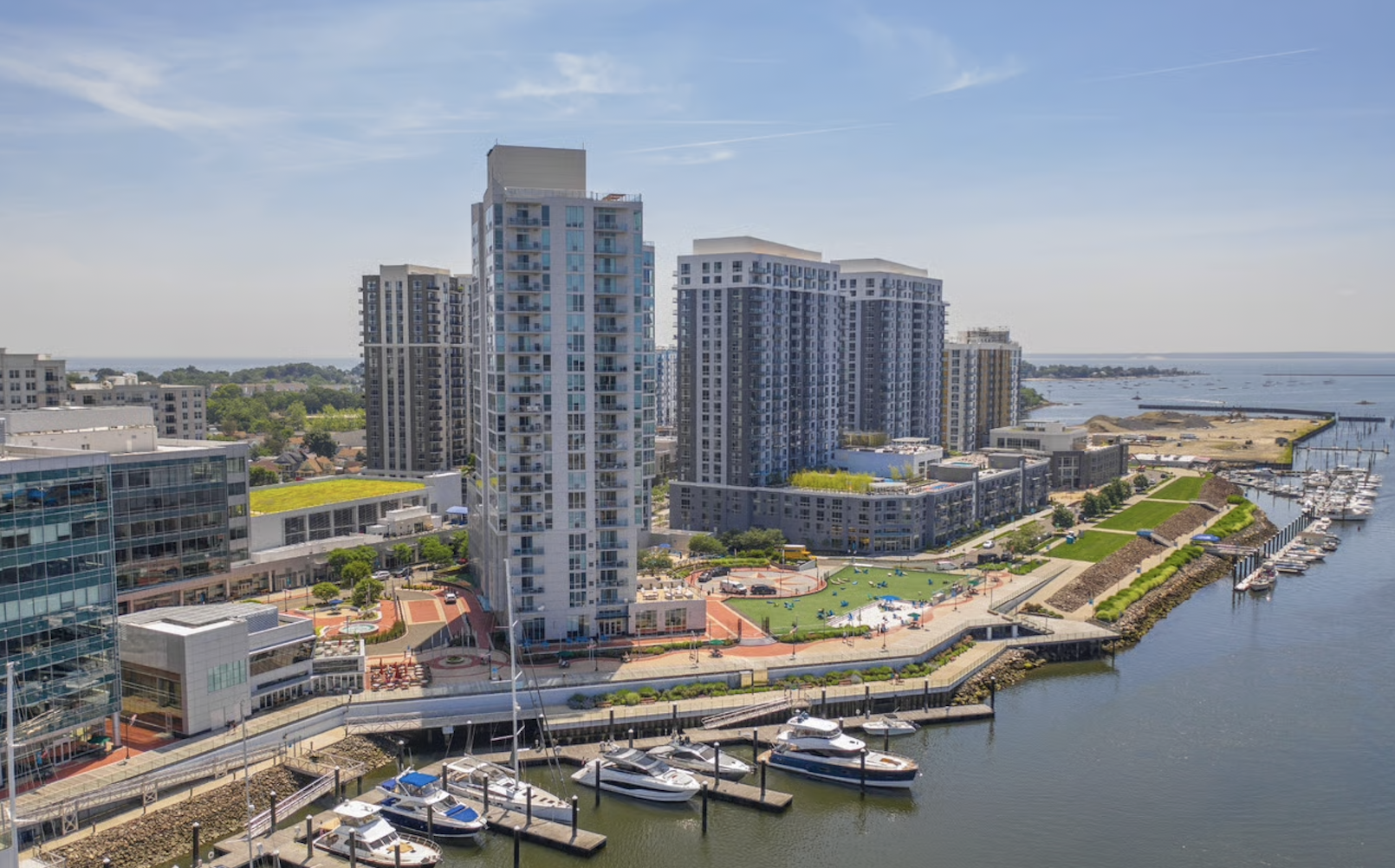 A marina with boats docked along water in front of tall modern apartment buildings and a park area with playground equipment.