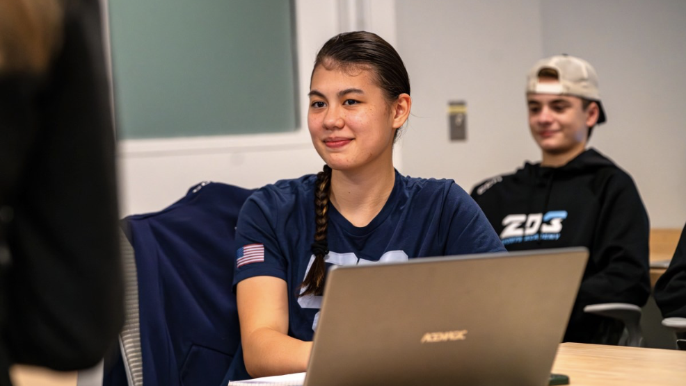 A young woman with a braid, wearing a navy sports shirt with an American flag patch, sits at a table with a laptop, smiling. Behind her, a young man in a black hoodie and cap also sits, smiling in a classroom or meeting room setting.