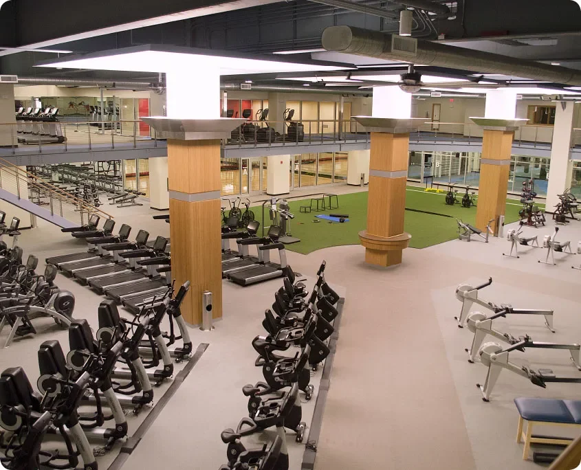 Modern gym with cardio machines, weight equipment, and workout area, featuring large columns and glass walls.