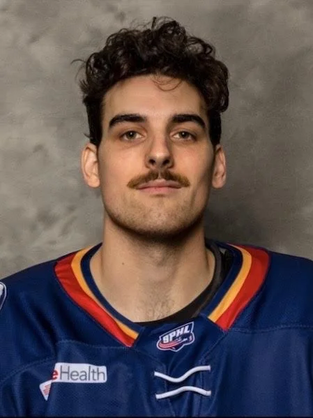 Young man with curly dark hair and a mustache wearing a blue hockey jersey with red, yellow, and blue accents, standing against a gray background.