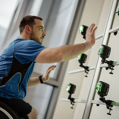 A man in a wheelchair reaching towards a row of electronic devices mounted on a wall at an indoor location.