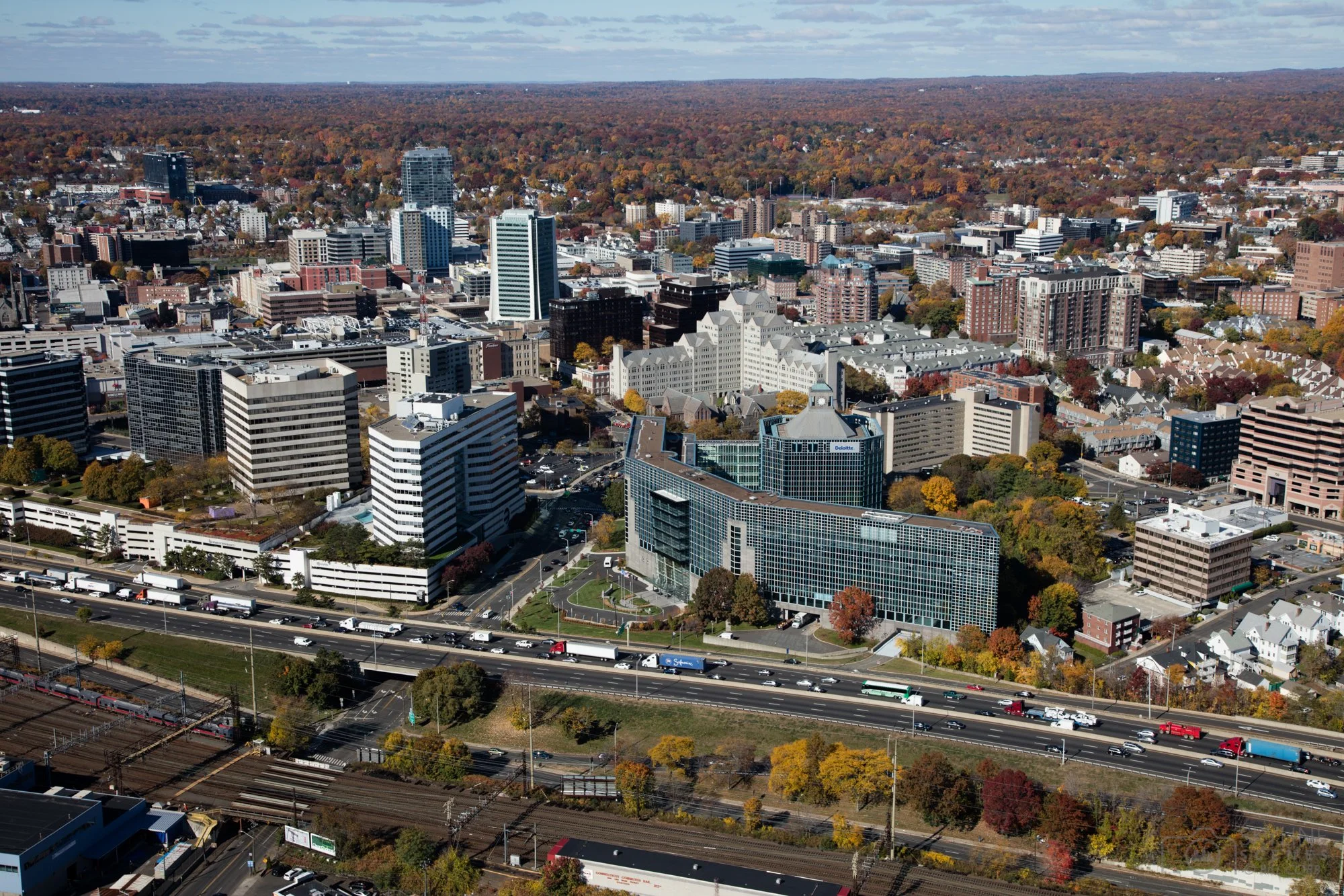 Aerial view of a city skyline with tall buildings, residential areas, highways, and train tracks, surrounded by a landscape of trees with autumn foliage.