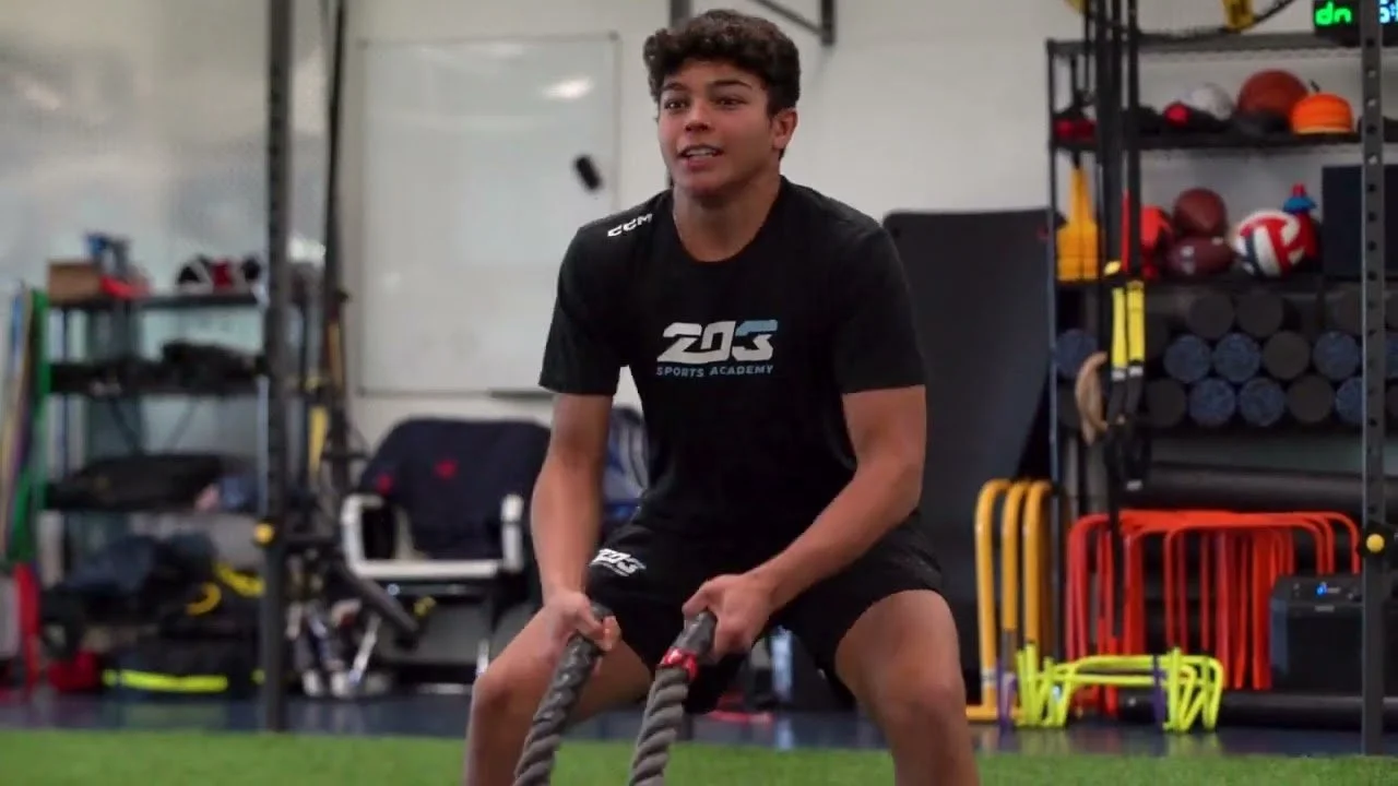 Teenage boy in black sports shirt and shorts training with battle ropes in a gym with sports equipment and workout gear in the background.