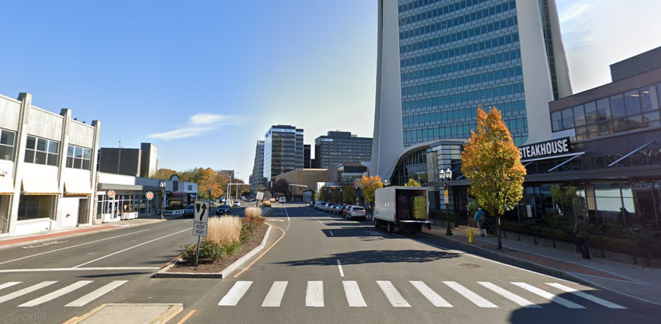 City street with modern buildings, parked cars, trees with autumn leaves, and a hotel with a sign indicating a steakhouse.