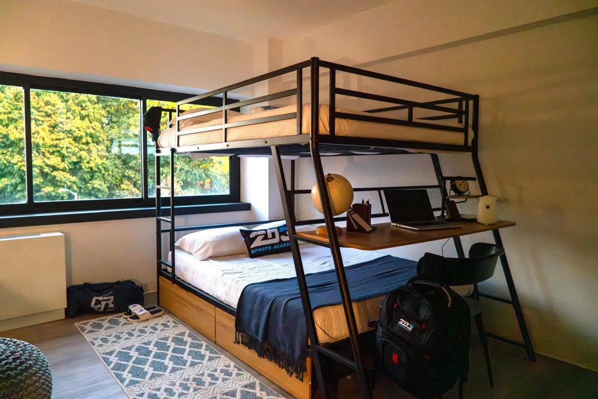 A modern bedroom with a loft bed and desk, large window showing trees outside, and various personal items including a backpack, laptop, and lamp.