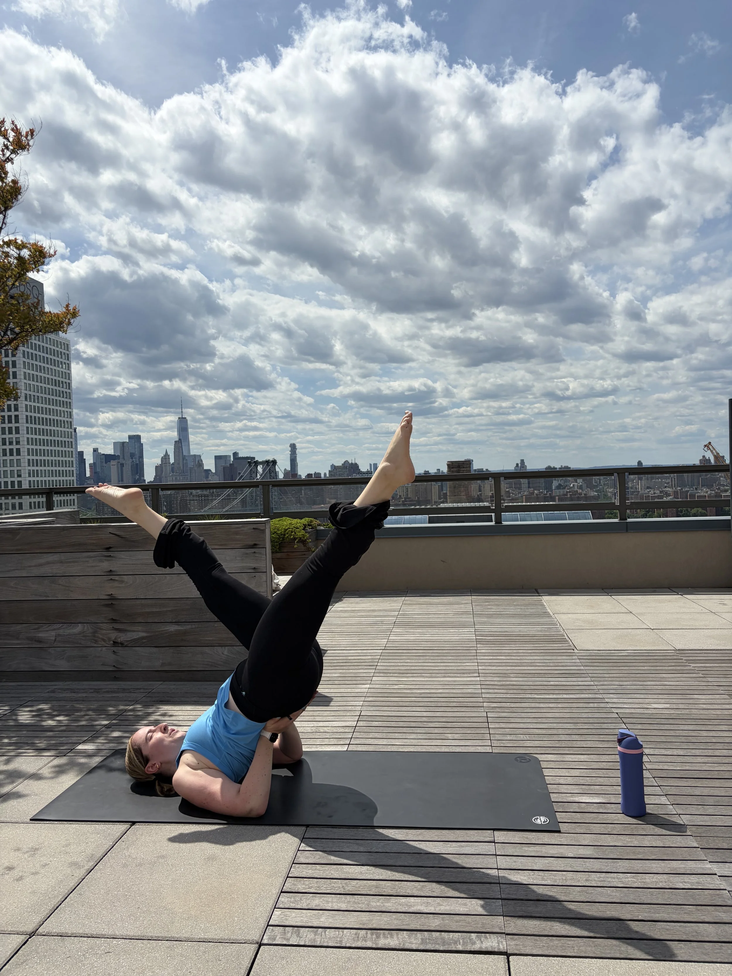 A woman performing a Pilates pose on a rooftop balcony with the New York City skyline and partly cloudy sky in the background.