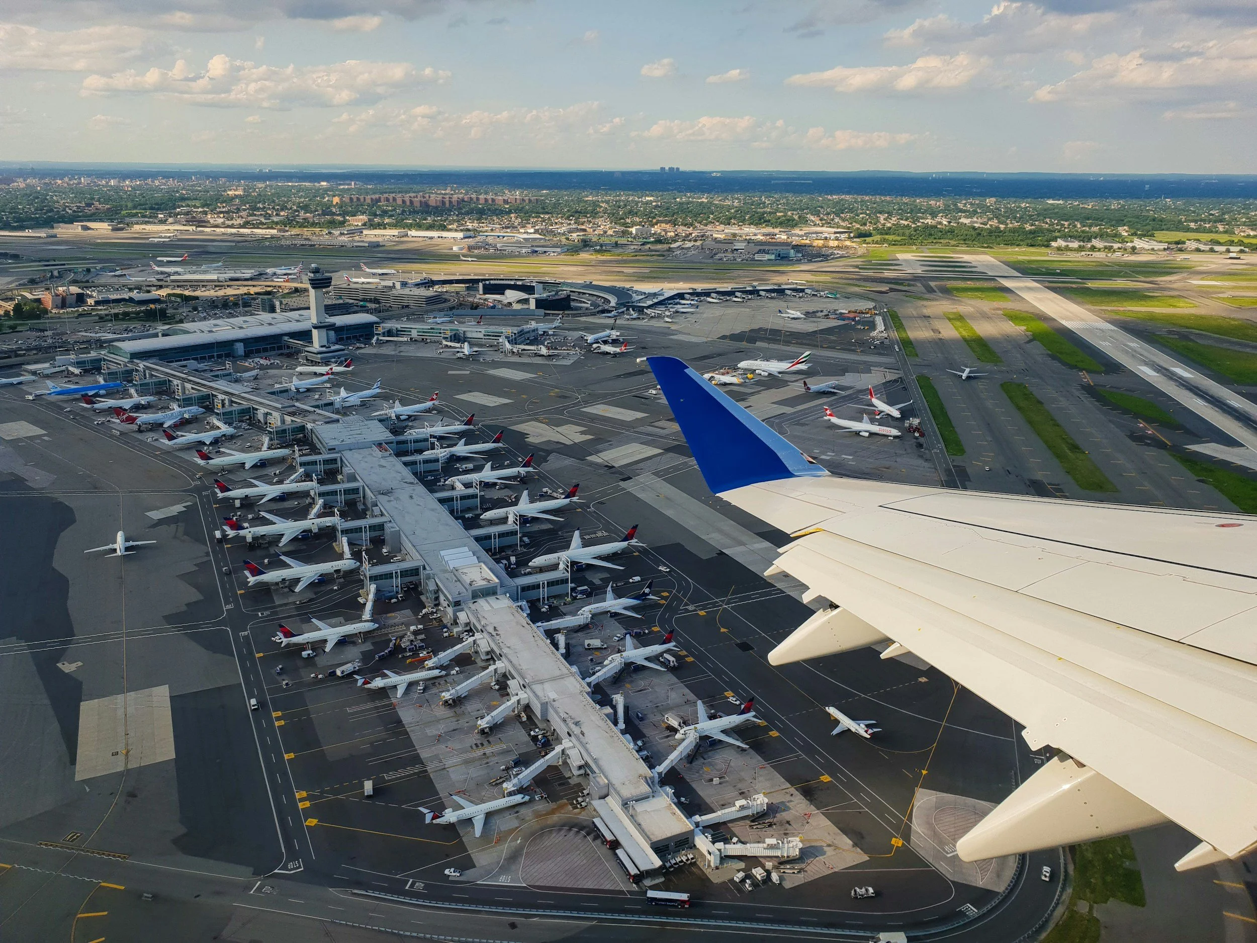 Vue aérienne d'un aéroport avec plusieurs avions stationnés aux portes et en piste, une tour de contrôle, des terminaux, et une ville en arrière-plan.