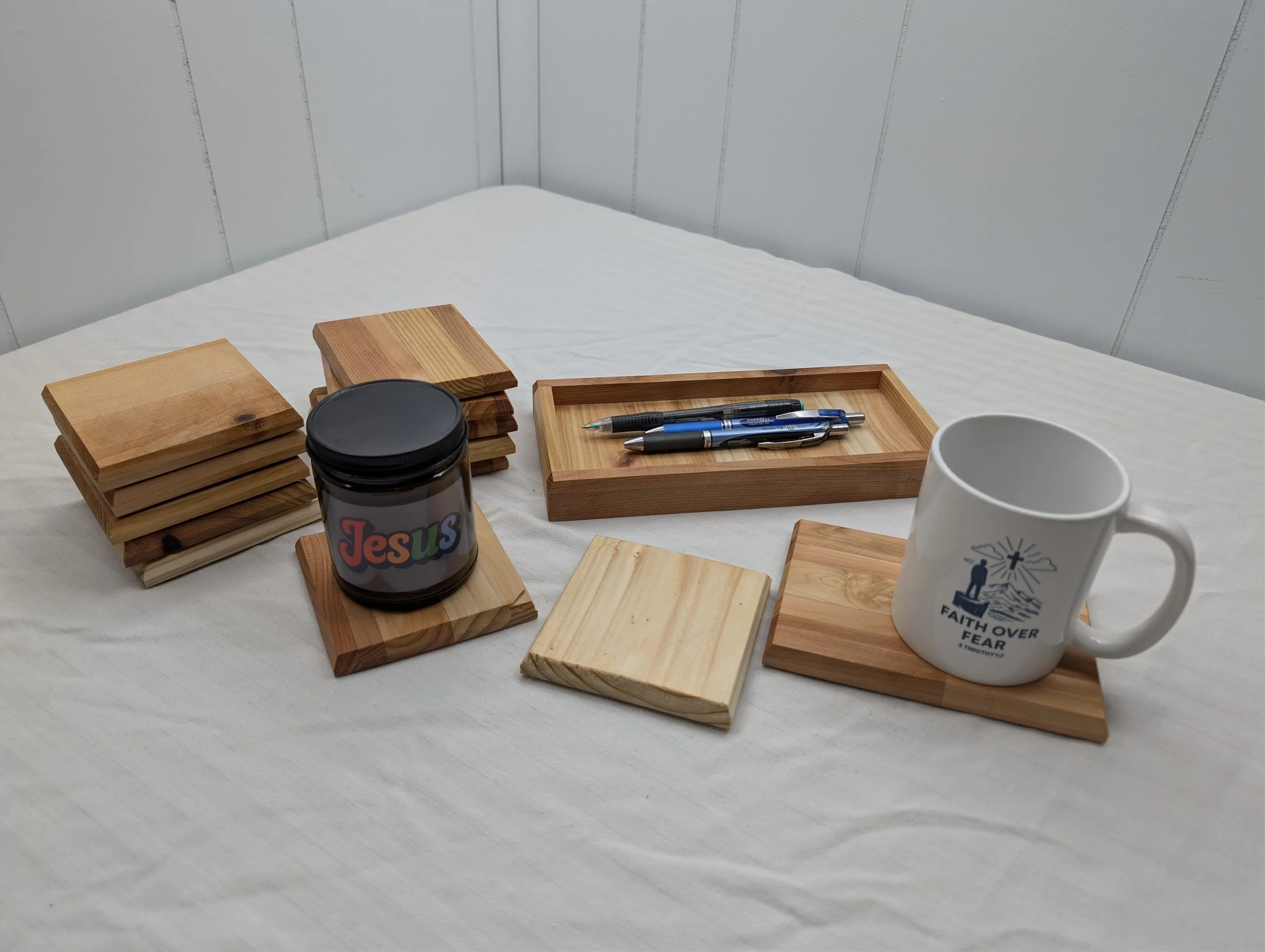 Arranged wooden coasters, pens, a black container labeled 'Jesus' with rainbow colors, a white mug with religious text, and a wooden tray on a white table, with a plain white wall in the background.